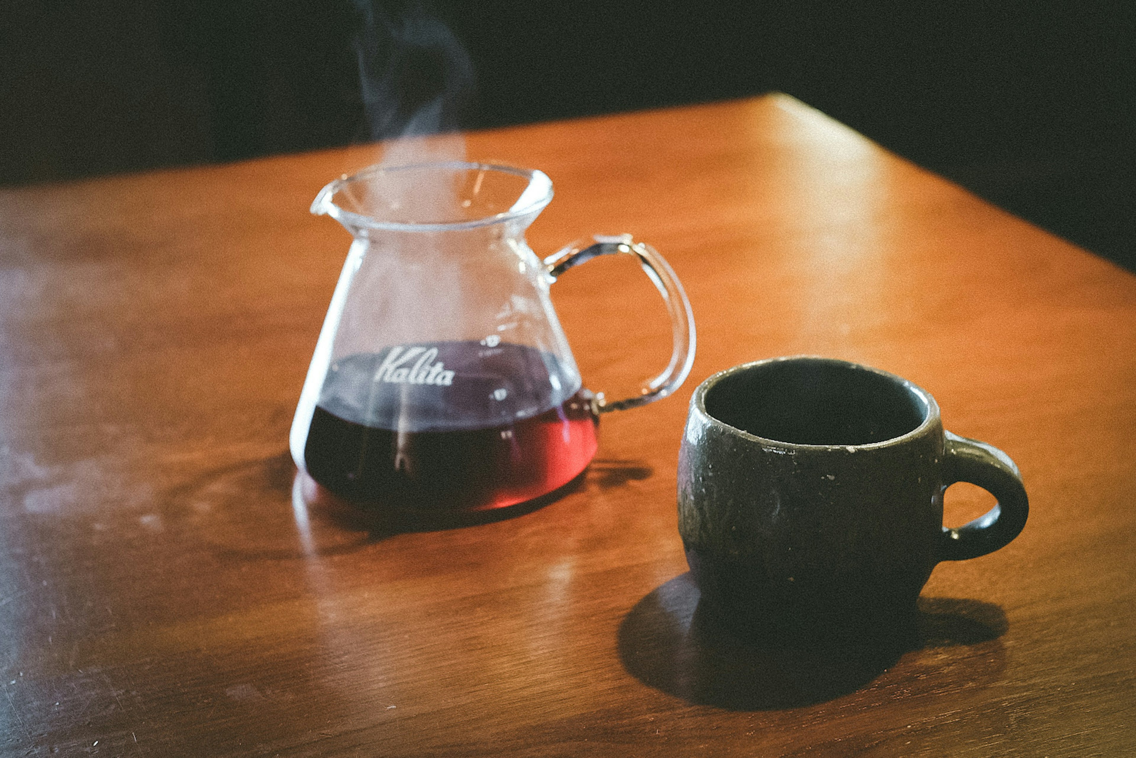 Una jarra transparente con té humeante y una taza negra sobre una mesa de madera