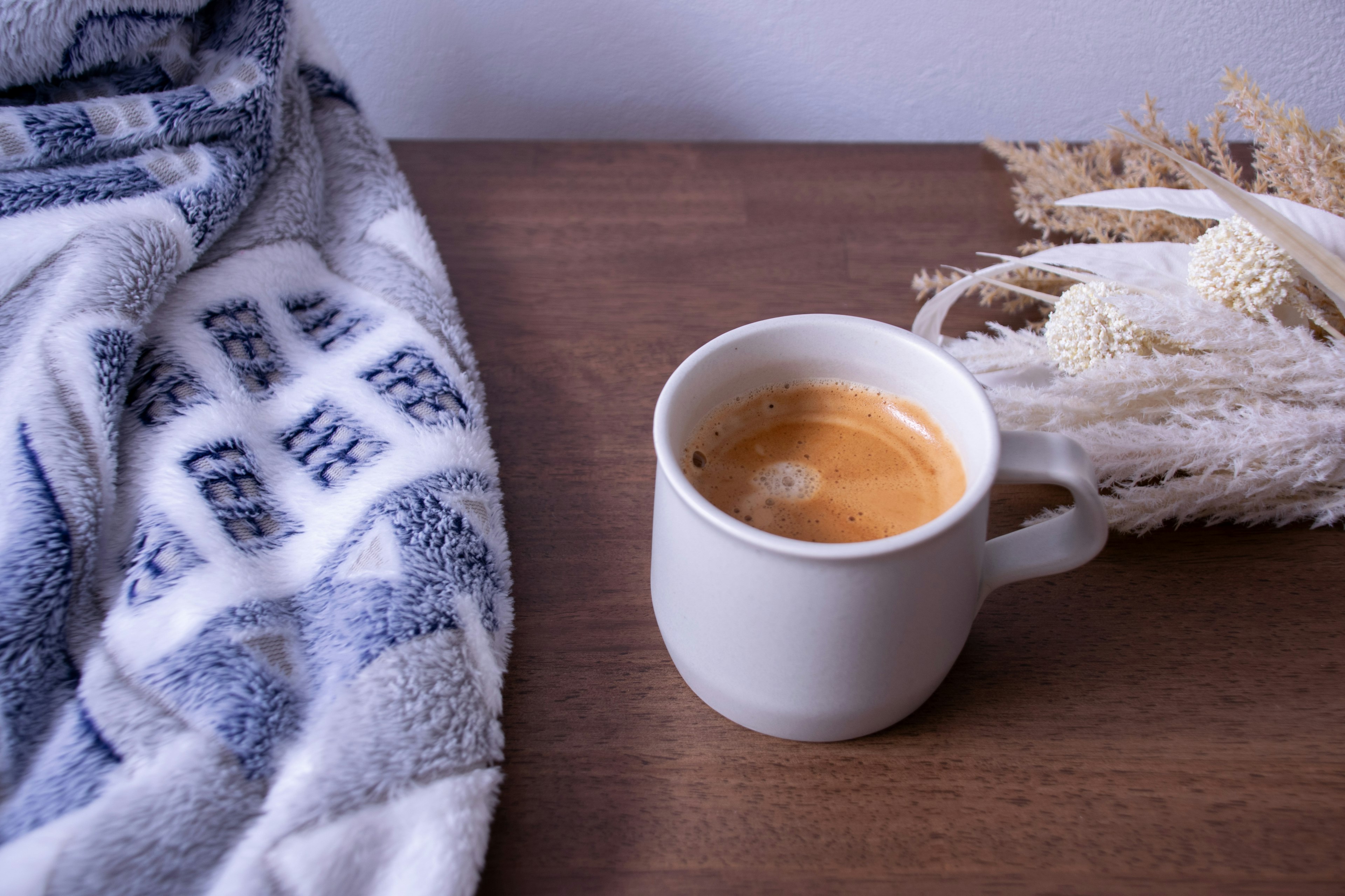 Eine gemütliche Szene mit einer weißen Espressotasse neben einer weichen Decke und getrockneten Blumen auf einem Holztisch