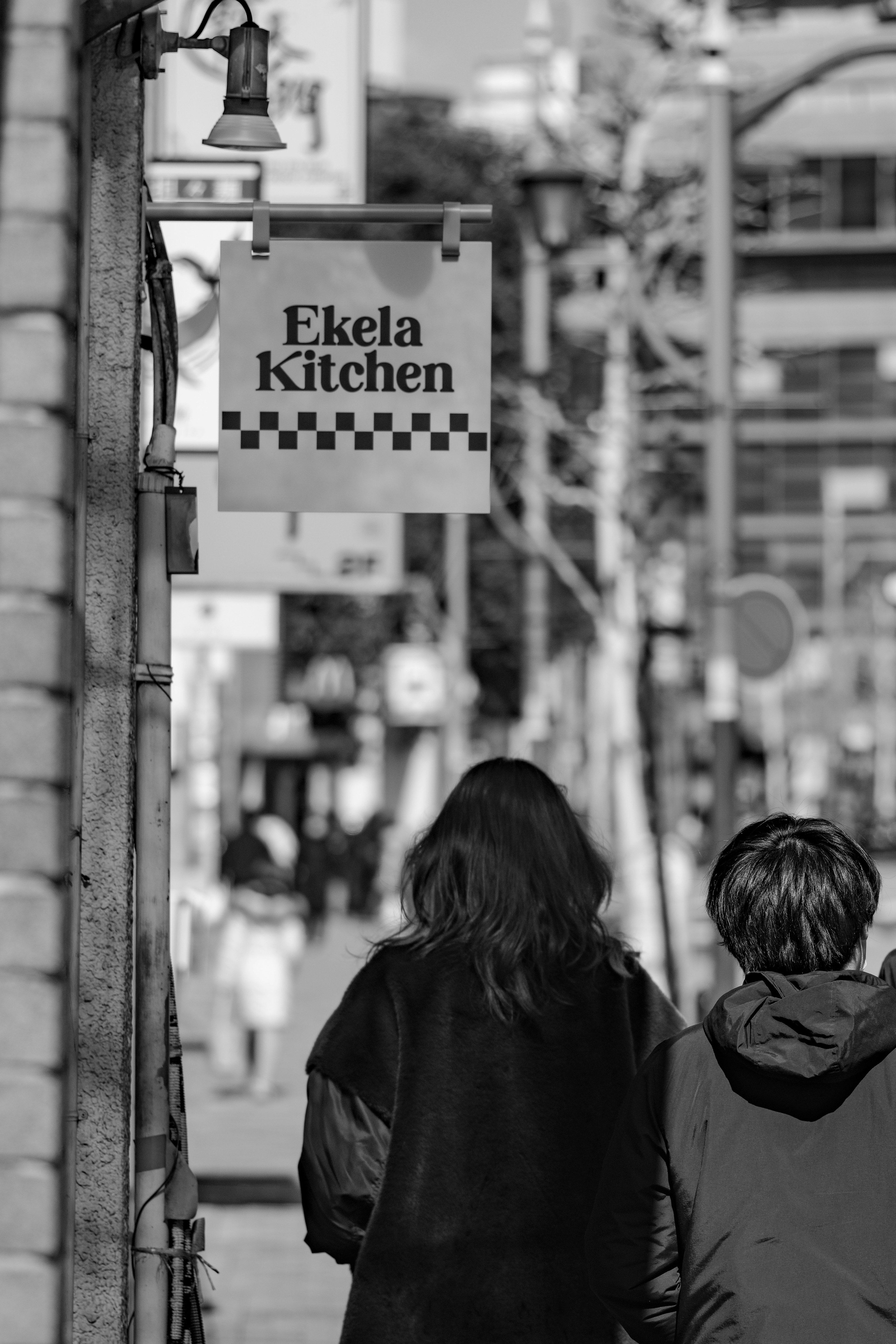 Two people walking away from the camera in a street scene featuring an Ekela Kitchen sign