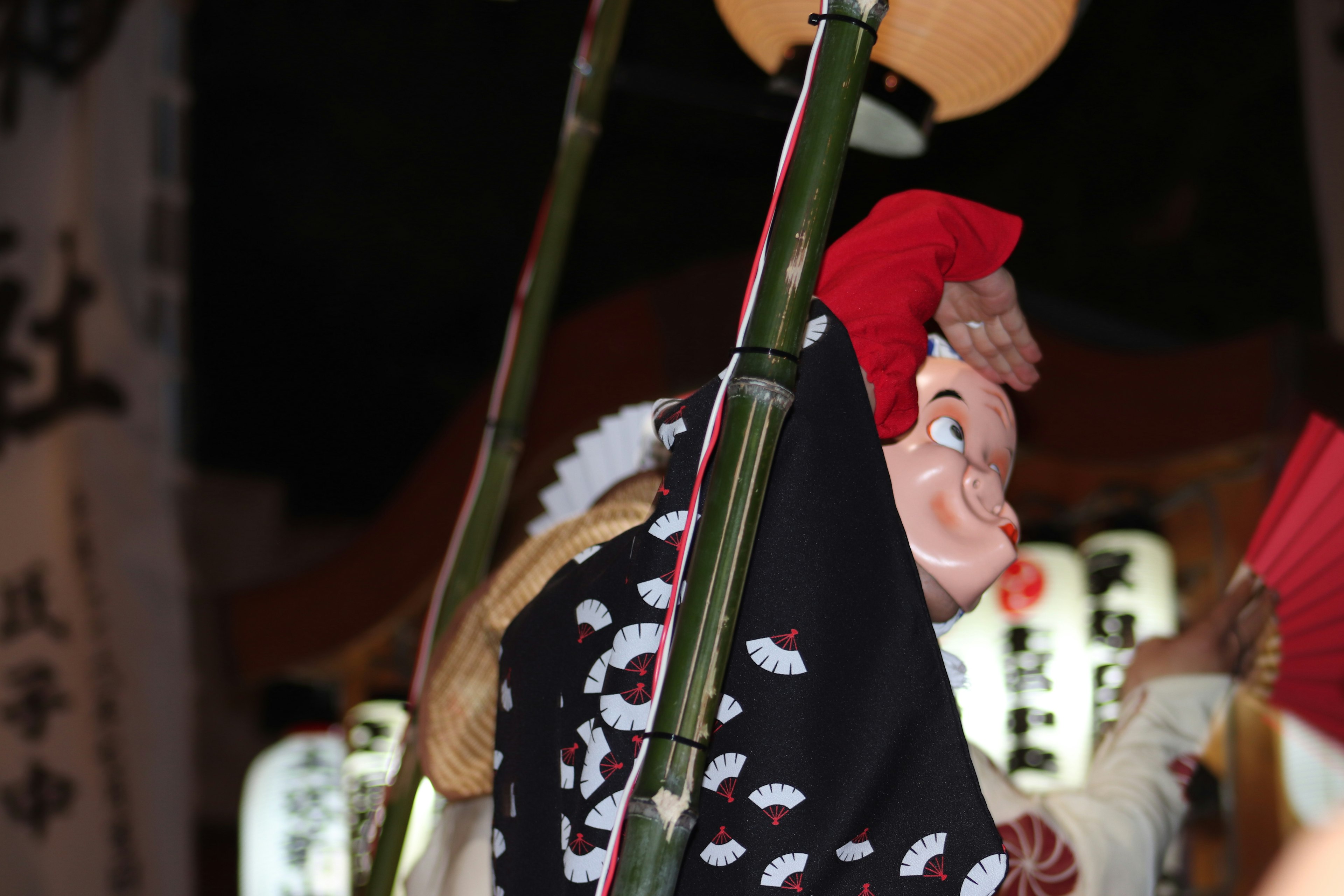 Sebuah boneka dengan sarung tangan merah menggantung dari tiang bambu dalam suasana festival malam