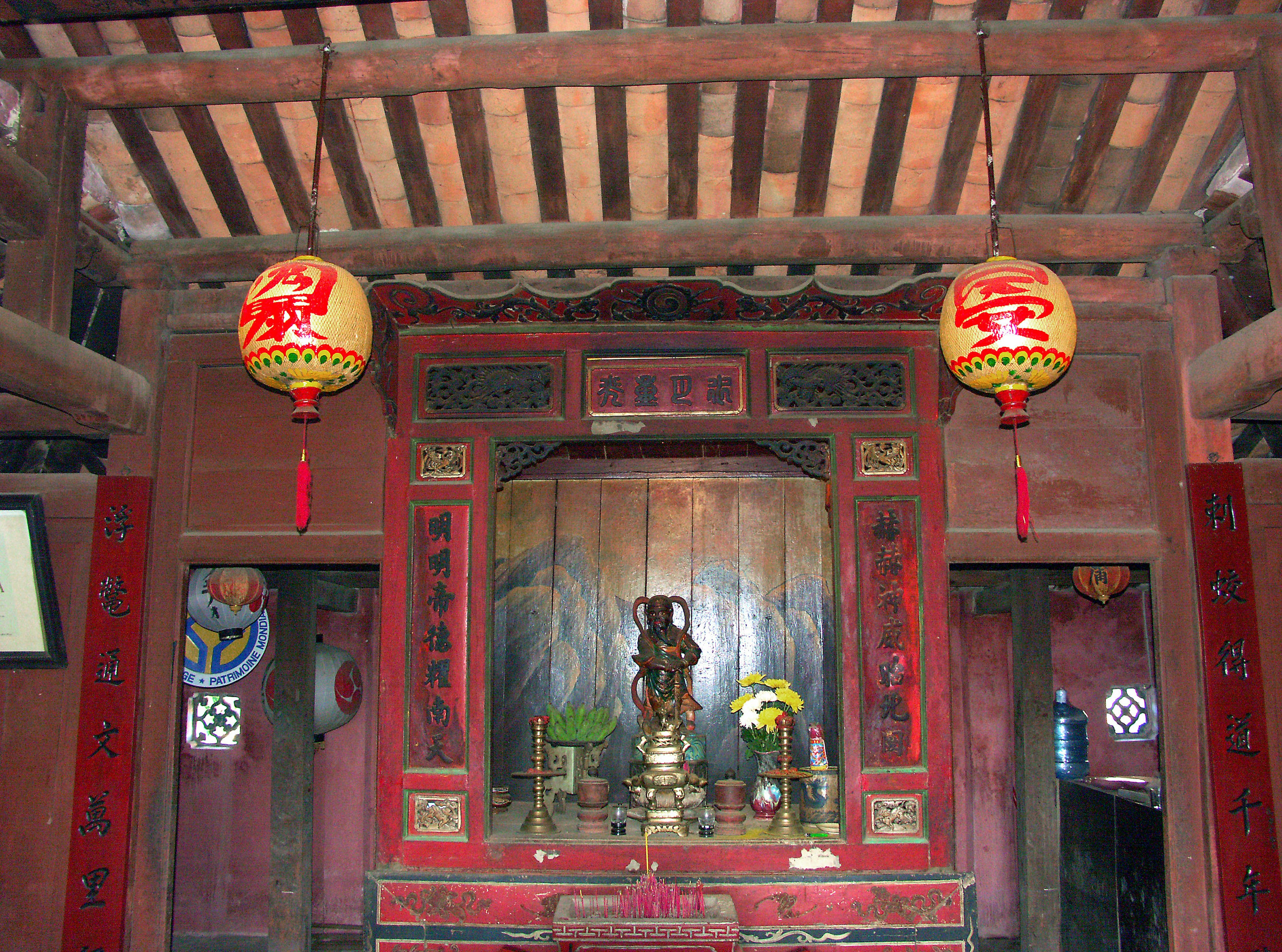 Interior de un templo tradicional con una estatua y flores en paredes rojas y vigas de madera