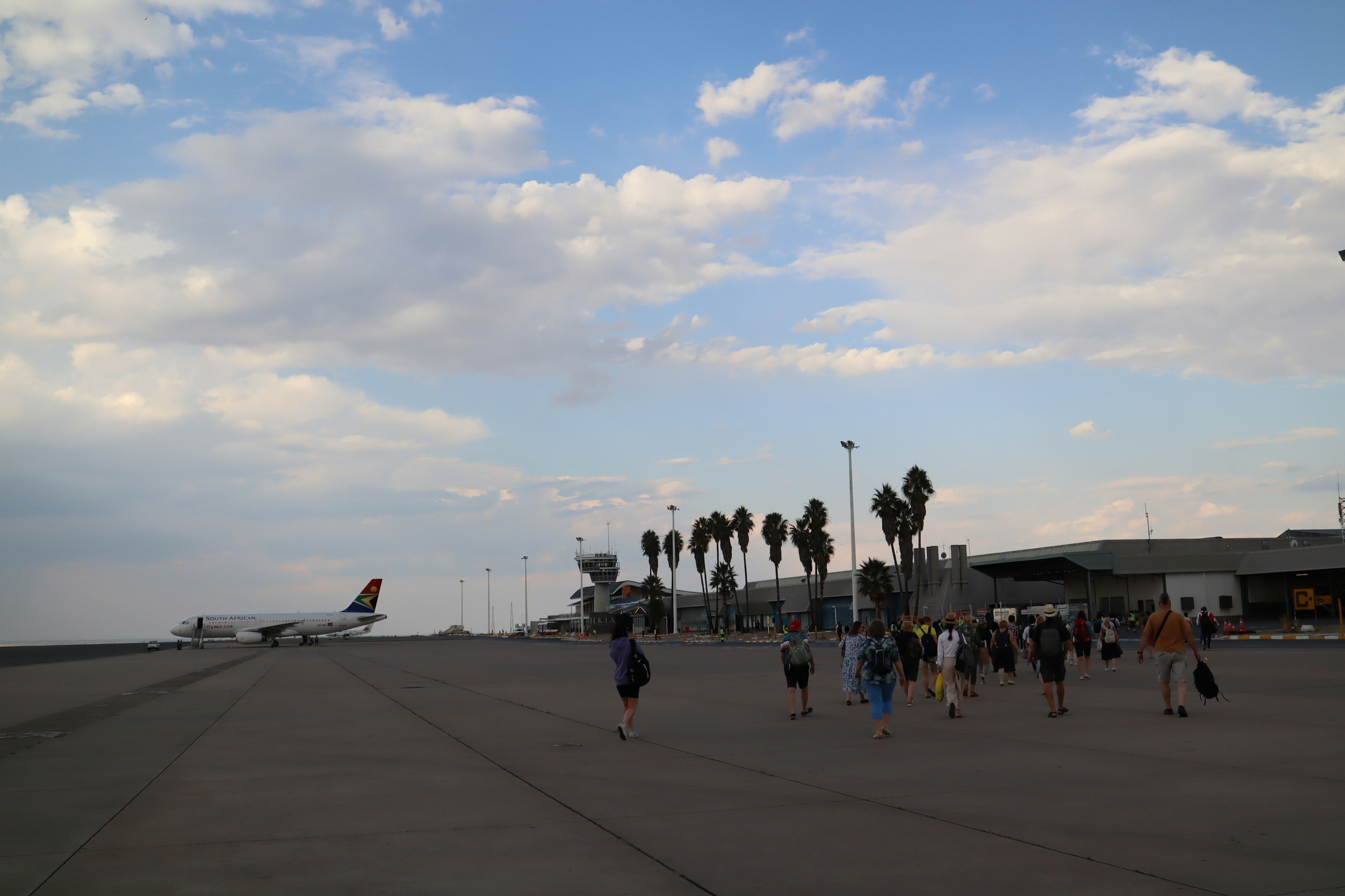 People walking on an airport runway with palm trees and a plane in the background