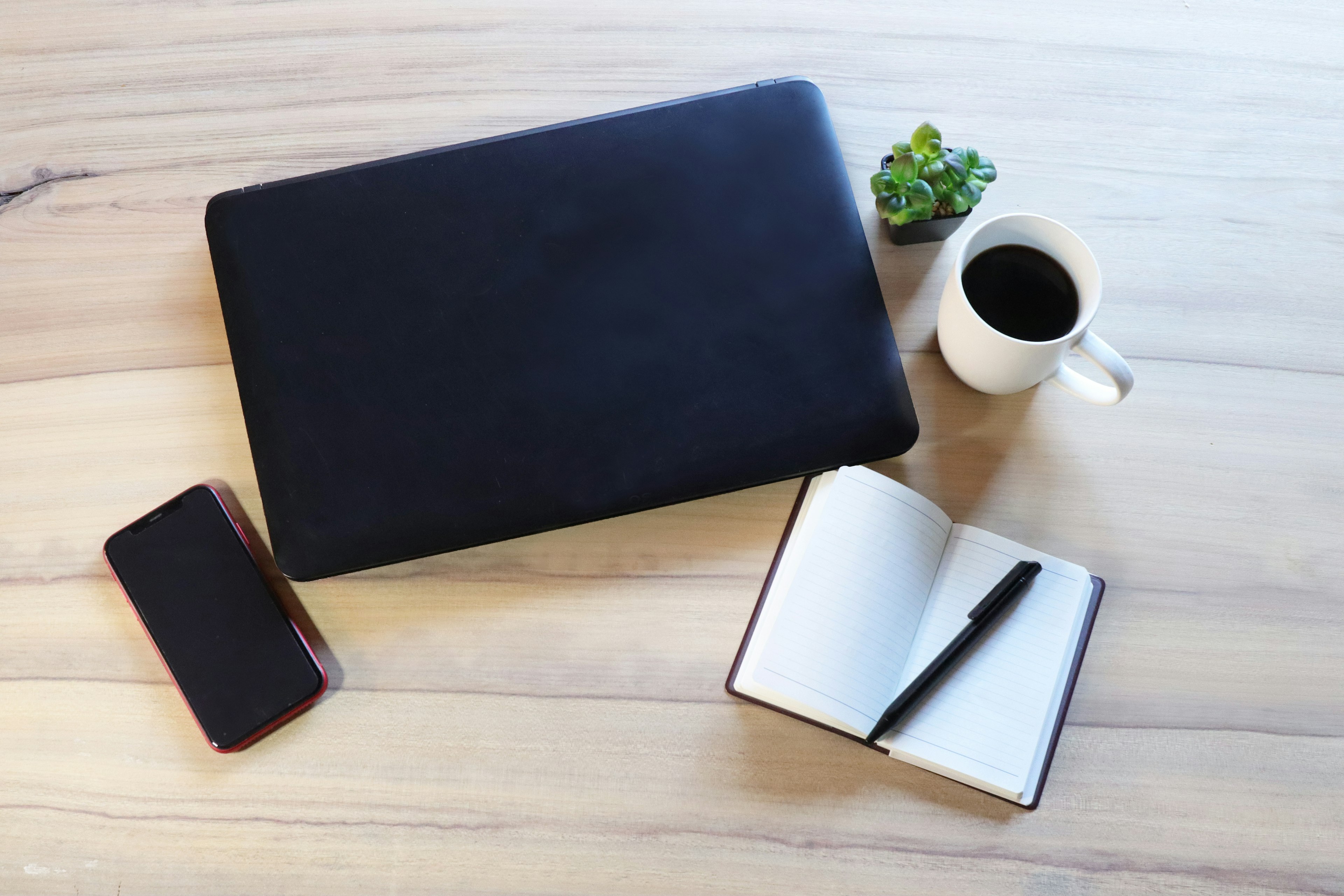 portátil negro y taza blanca sobre una mesa de madera con un cuaderno y un teléfono inteligente