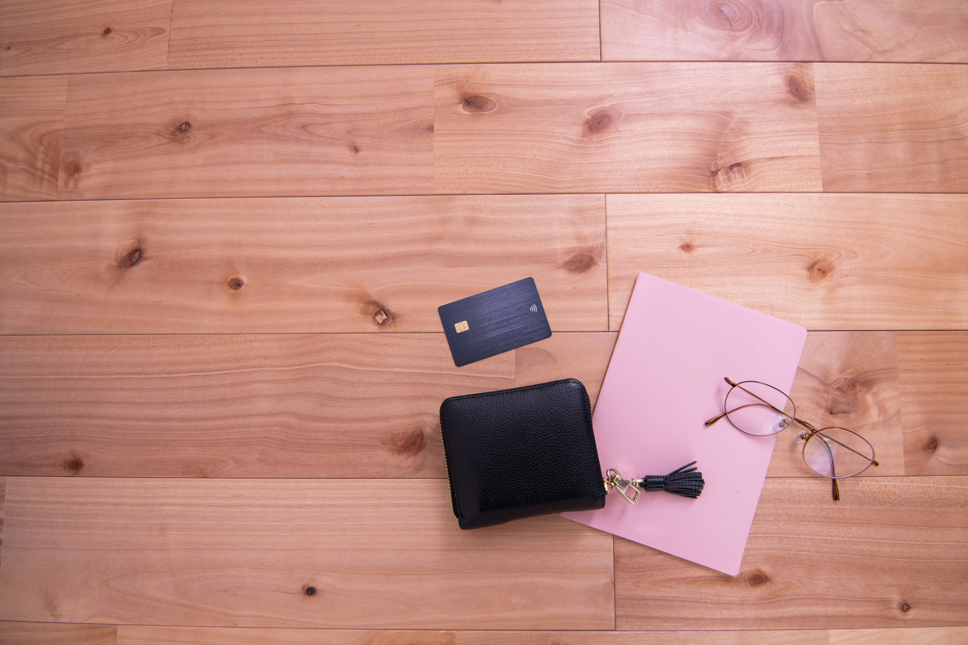 A black wallet and credit card next to a pink notebook and glasses on a wooden floor