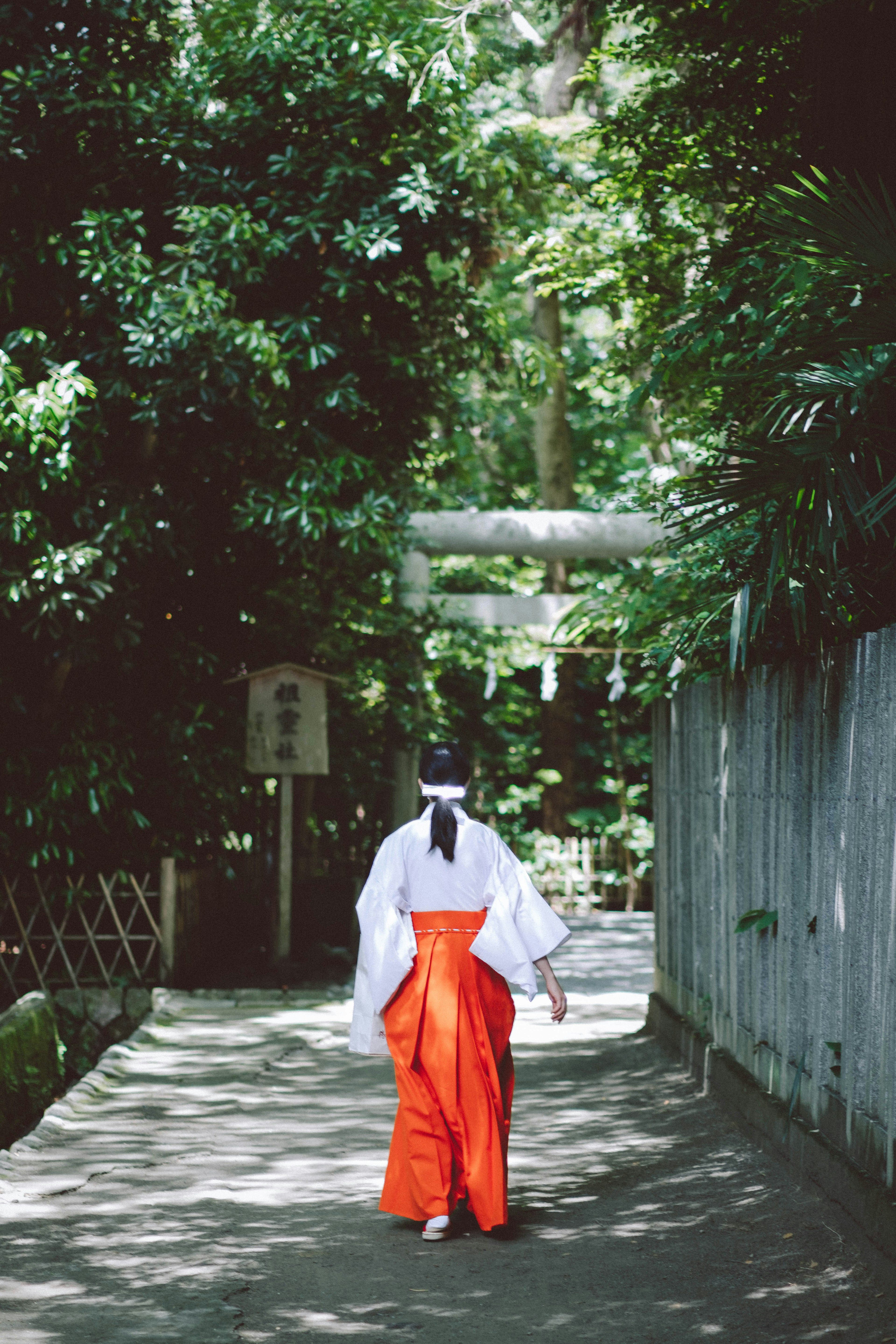 Una mujer en kimono blanco y hakama naranja caminando por un camino rodeado de árboles verdes