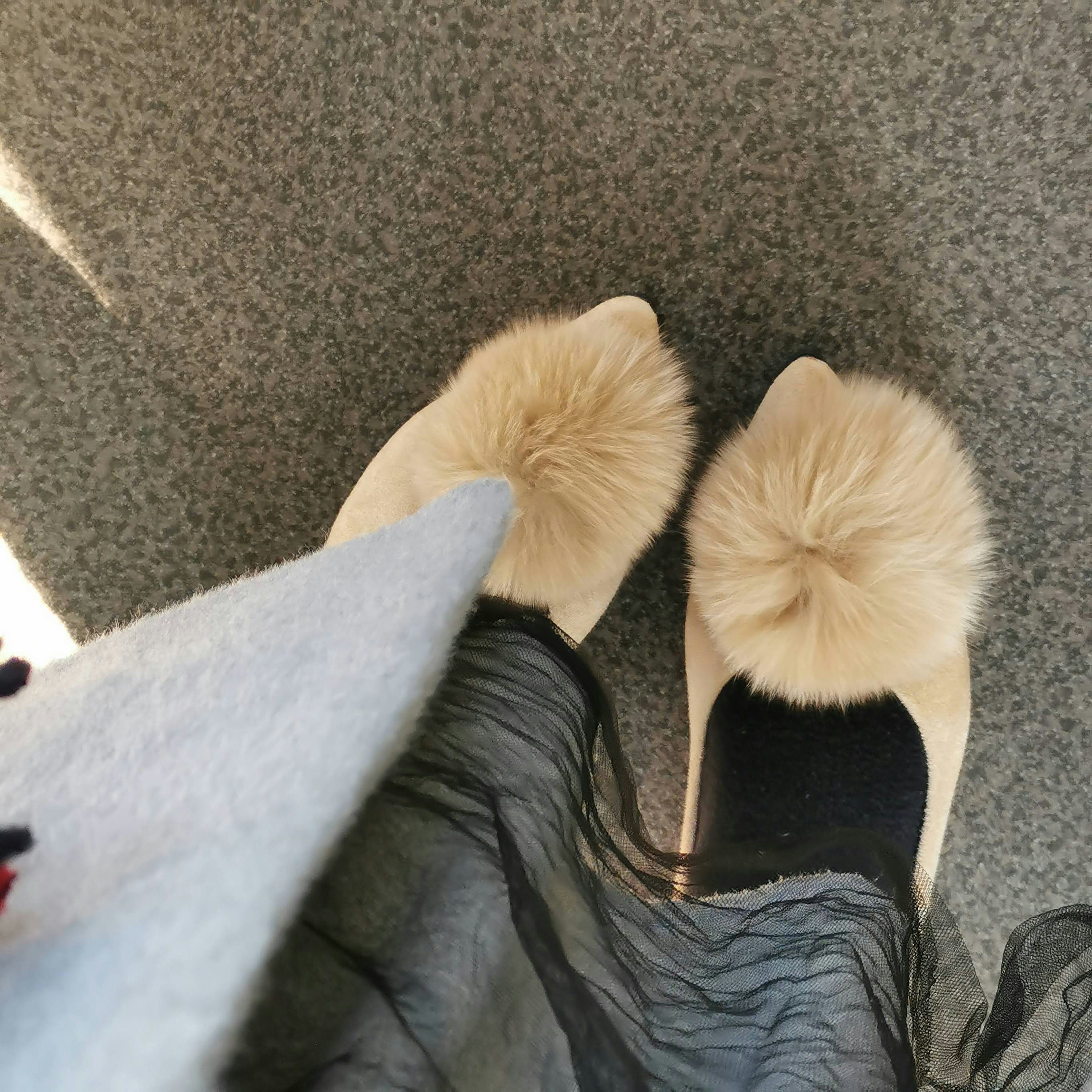 Feet in beige fur pom-pom slippers on a gray carpet