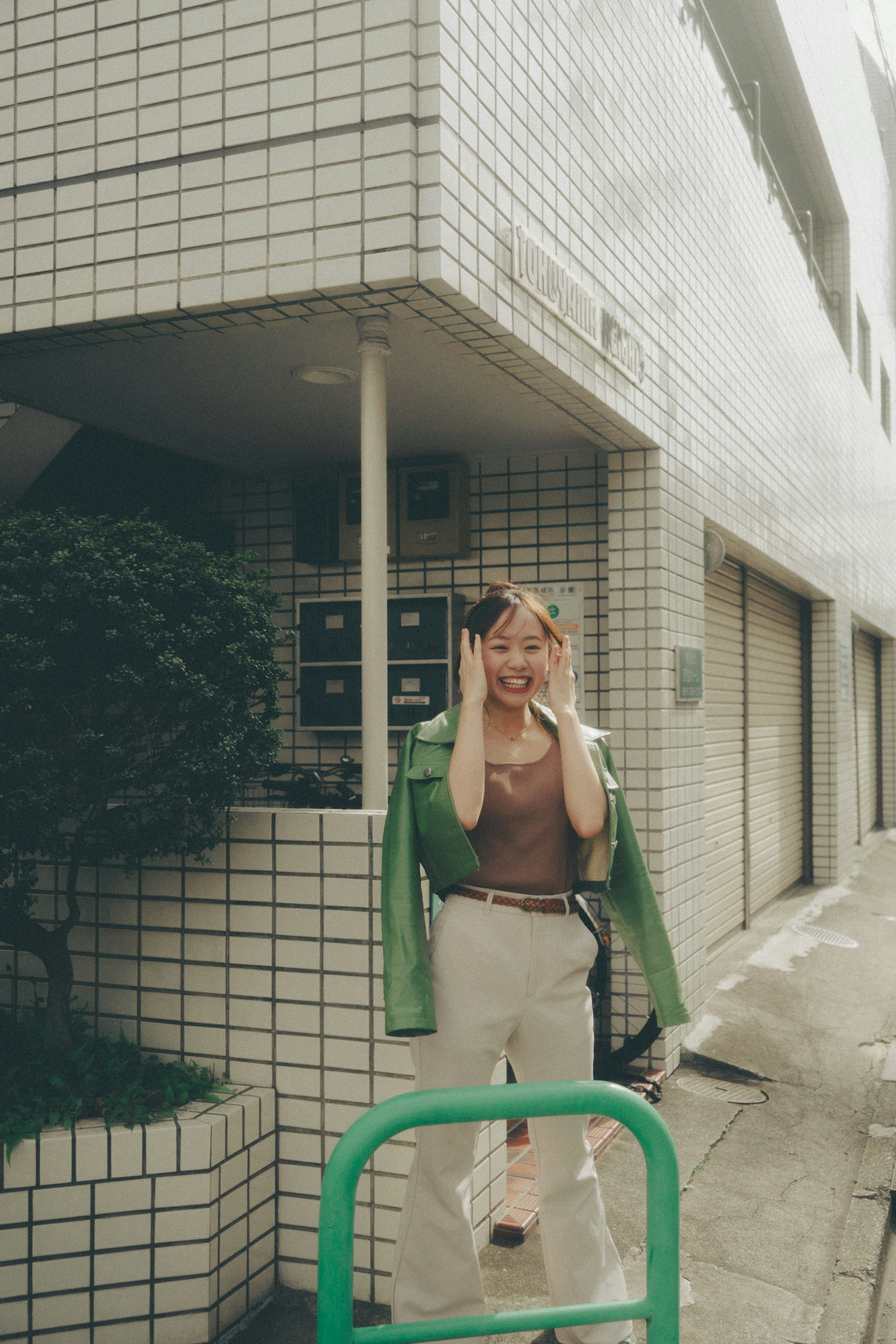 A smiling woman in a green jacket playfully touching her face in an urban setting