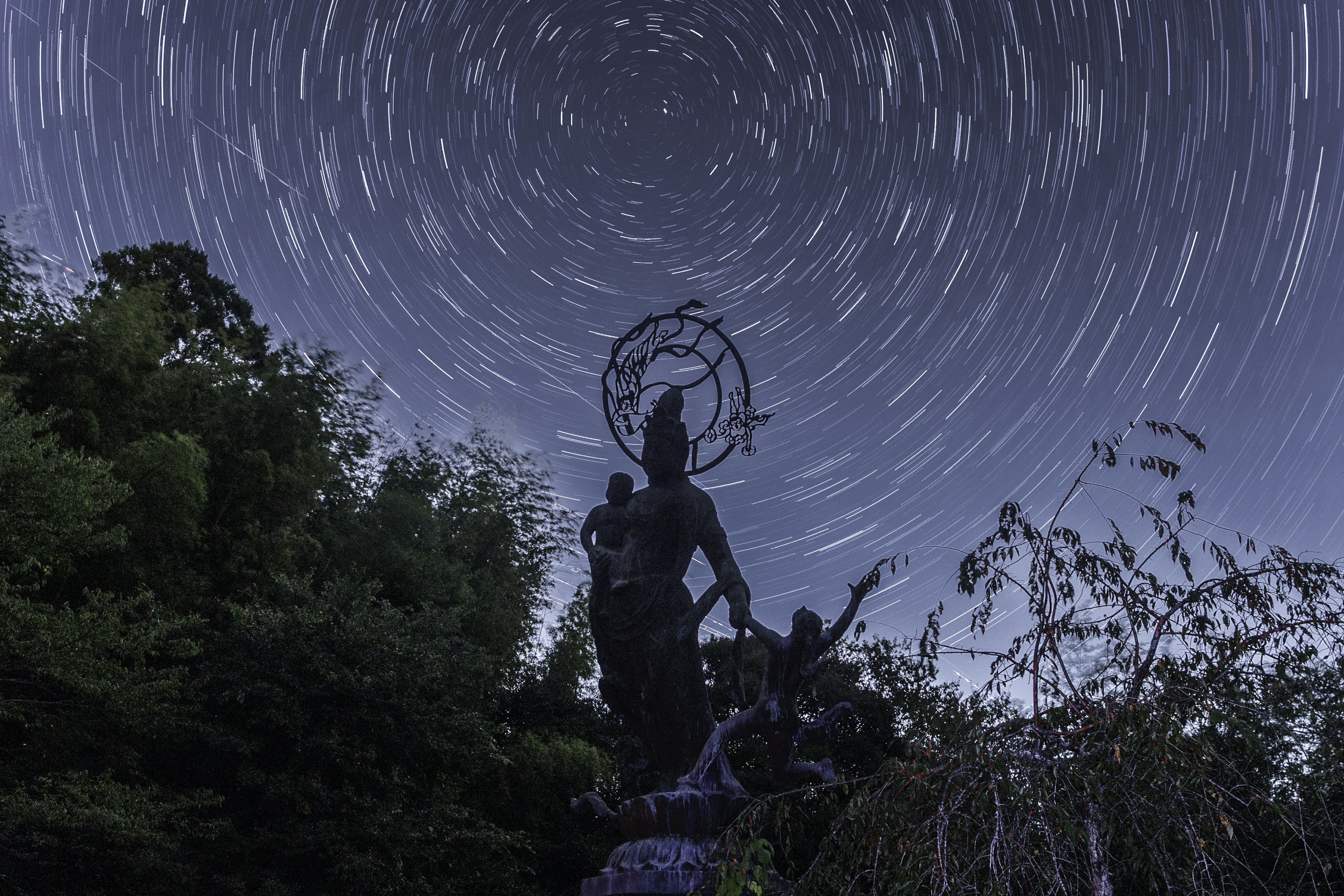 星の軌跡が見える夜空の下に立つ大きな像と人々のシルエット