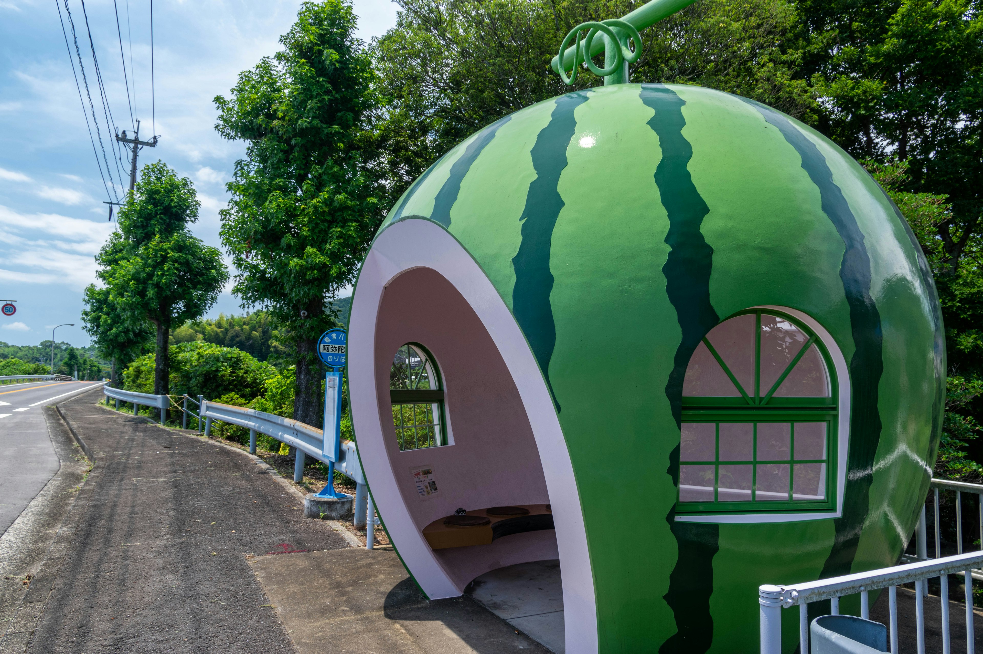 A watermelon-shaped bus stop located by the roadside surrounded by greenery