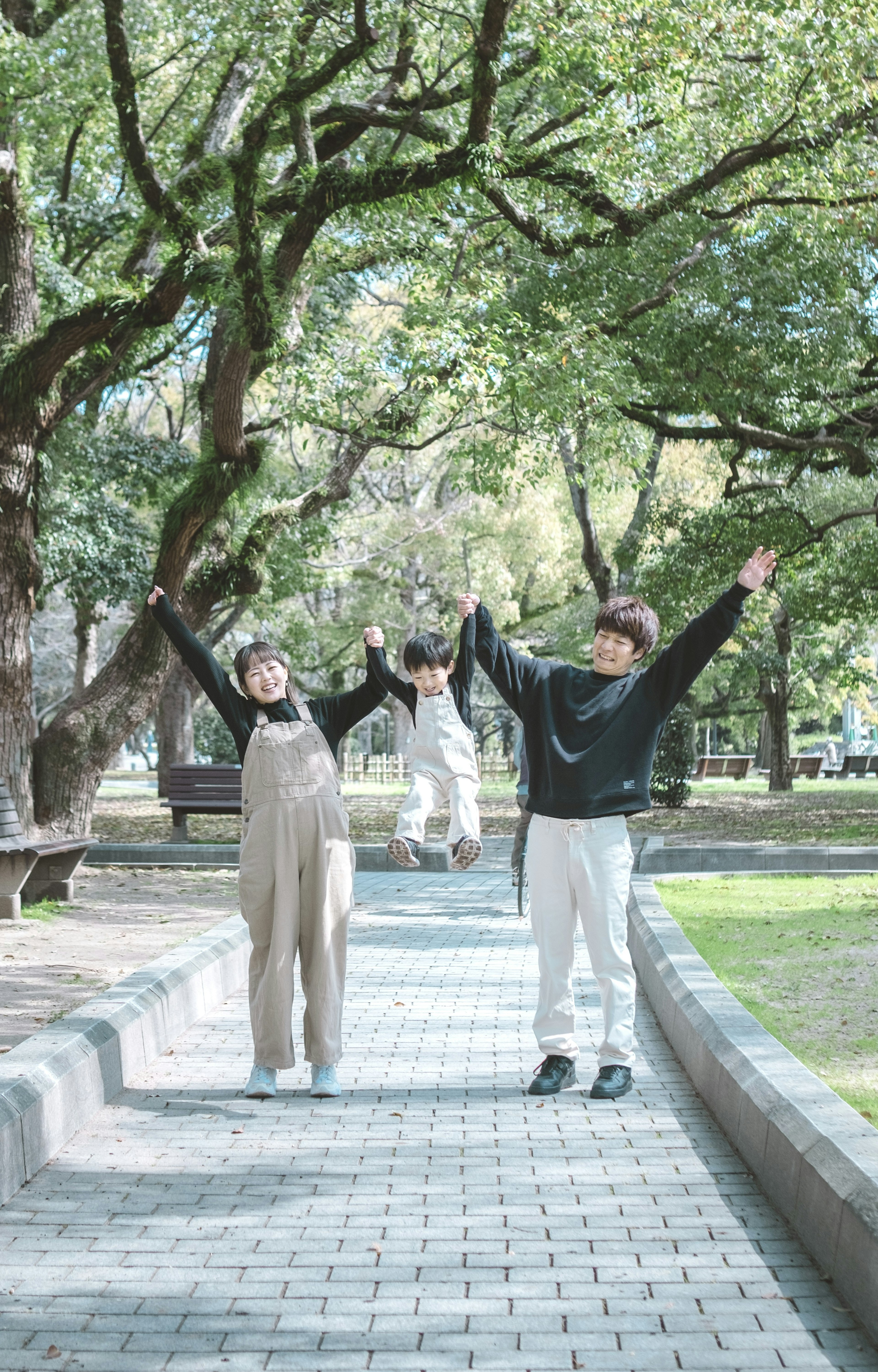 公園で楽しそうに手を挙げる二人の若者とその後ろにいる一人の人物 緑の木々に囲まれた歩道