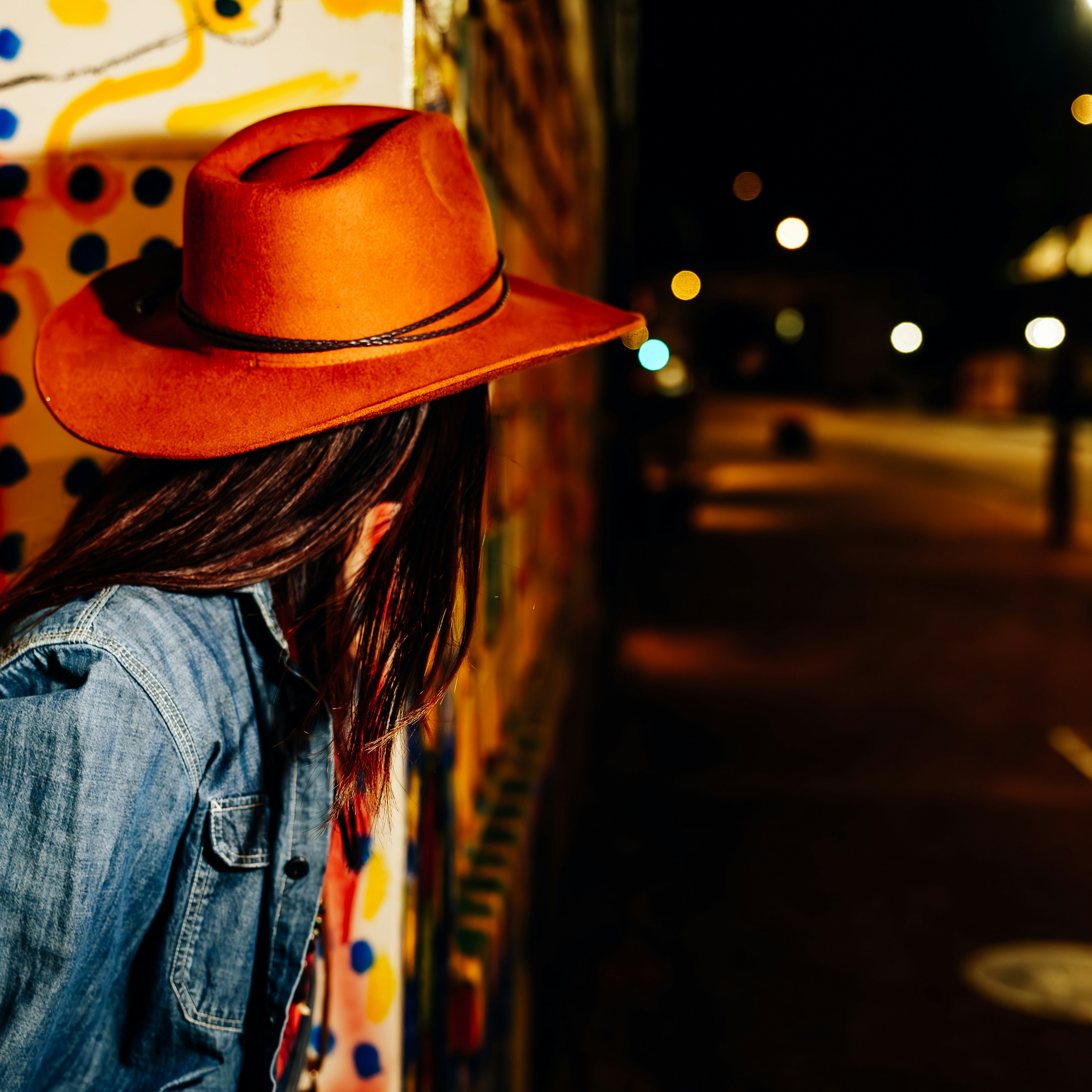 A person wearing an orange hat and denim jacket in a colorful urban setting at night