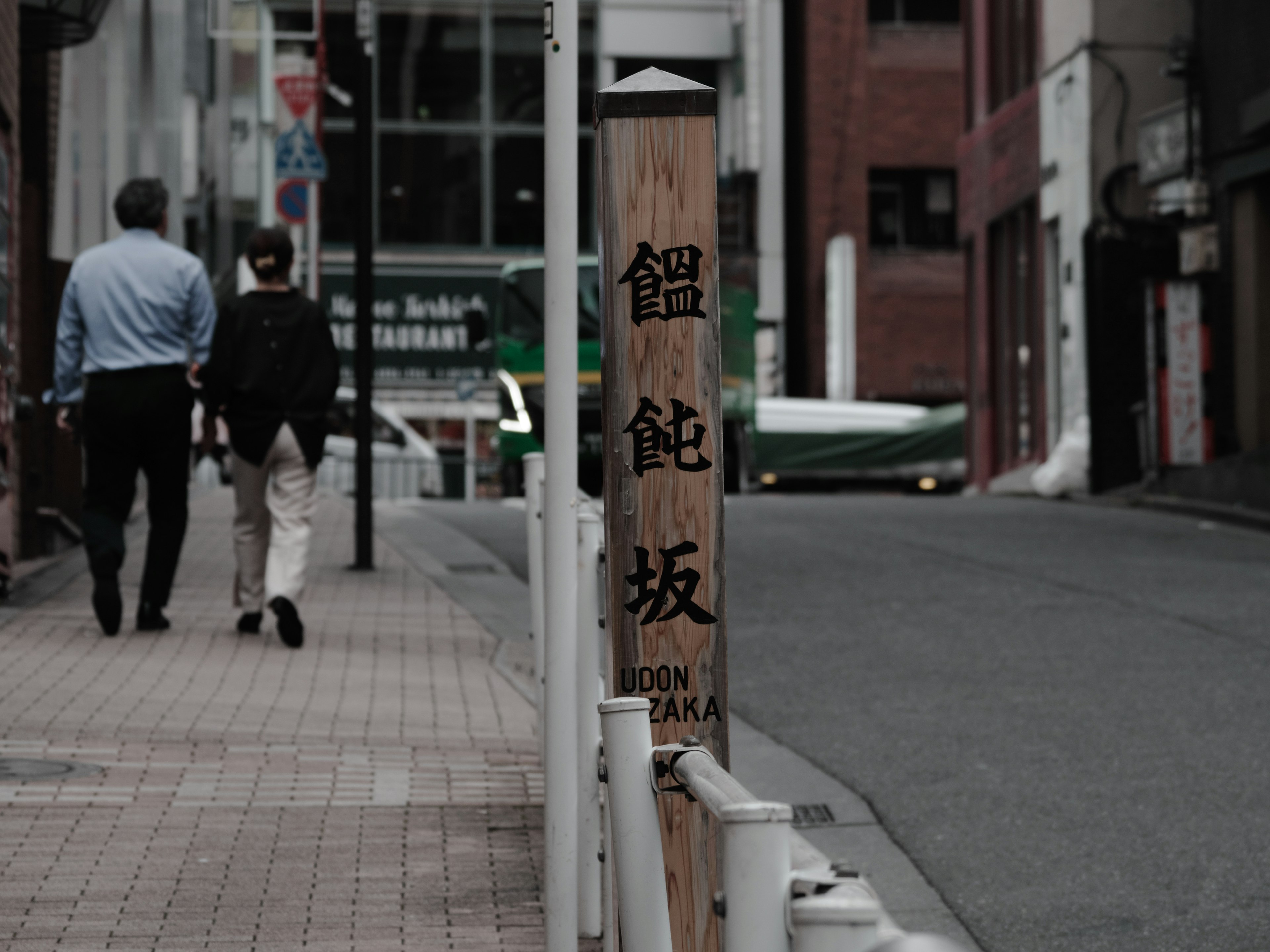 Two people walking on a street with a wooden sign visible