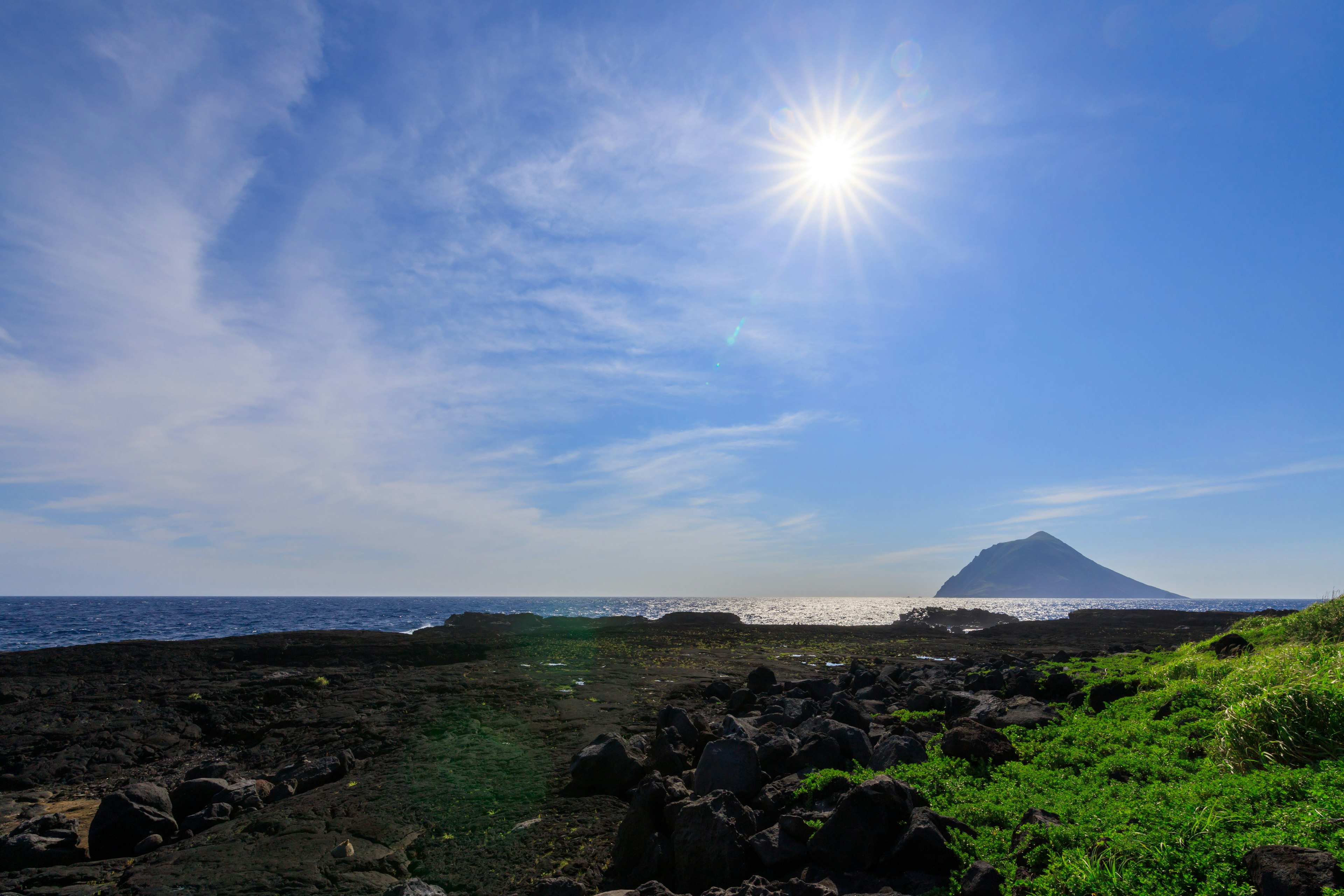 Paisaje costero con cielo azul brillante y sol rocas negras y hierba verde prominente montaña a lo lejos