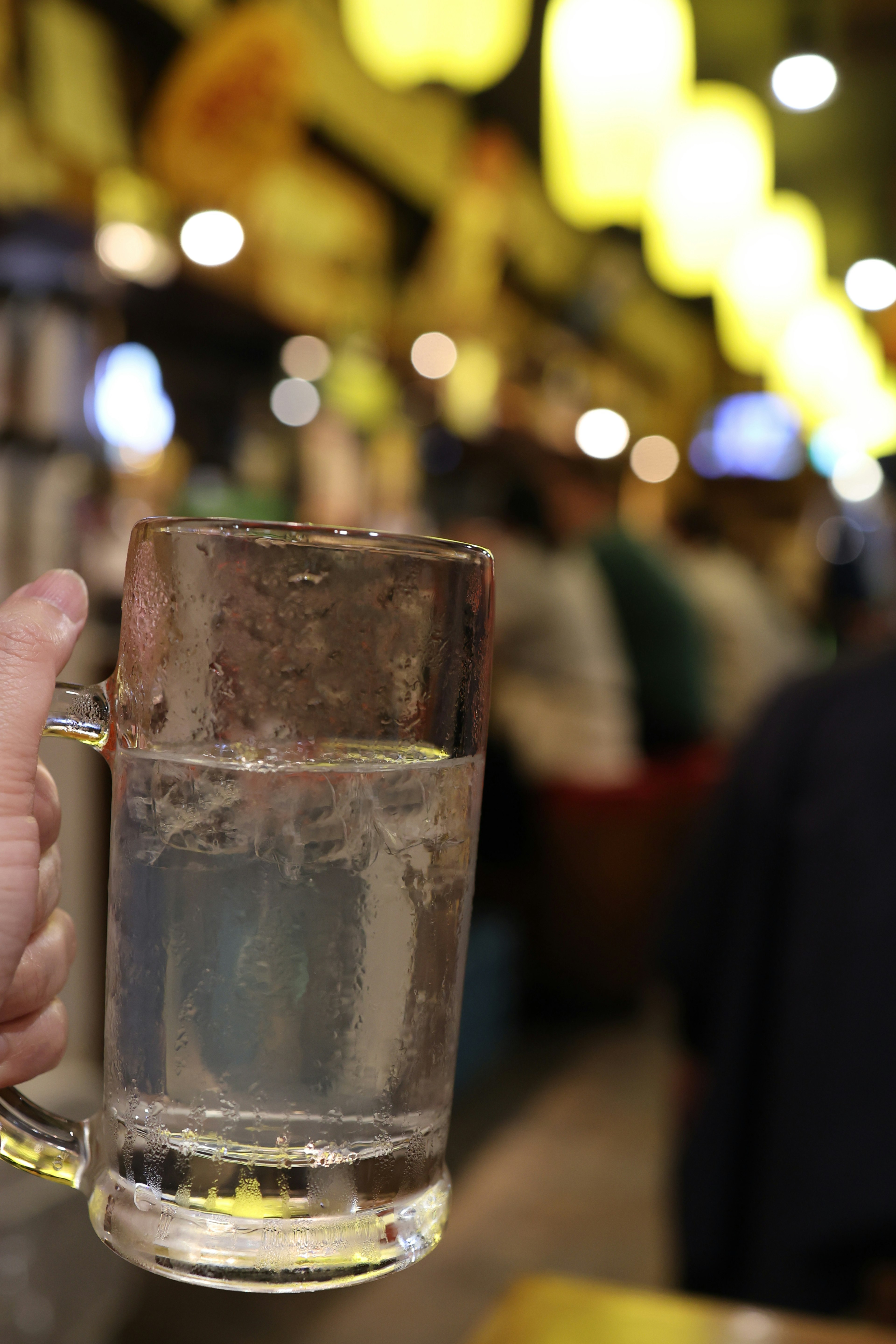 A hand holding a glass mug filled with clear water with blurred people in the background