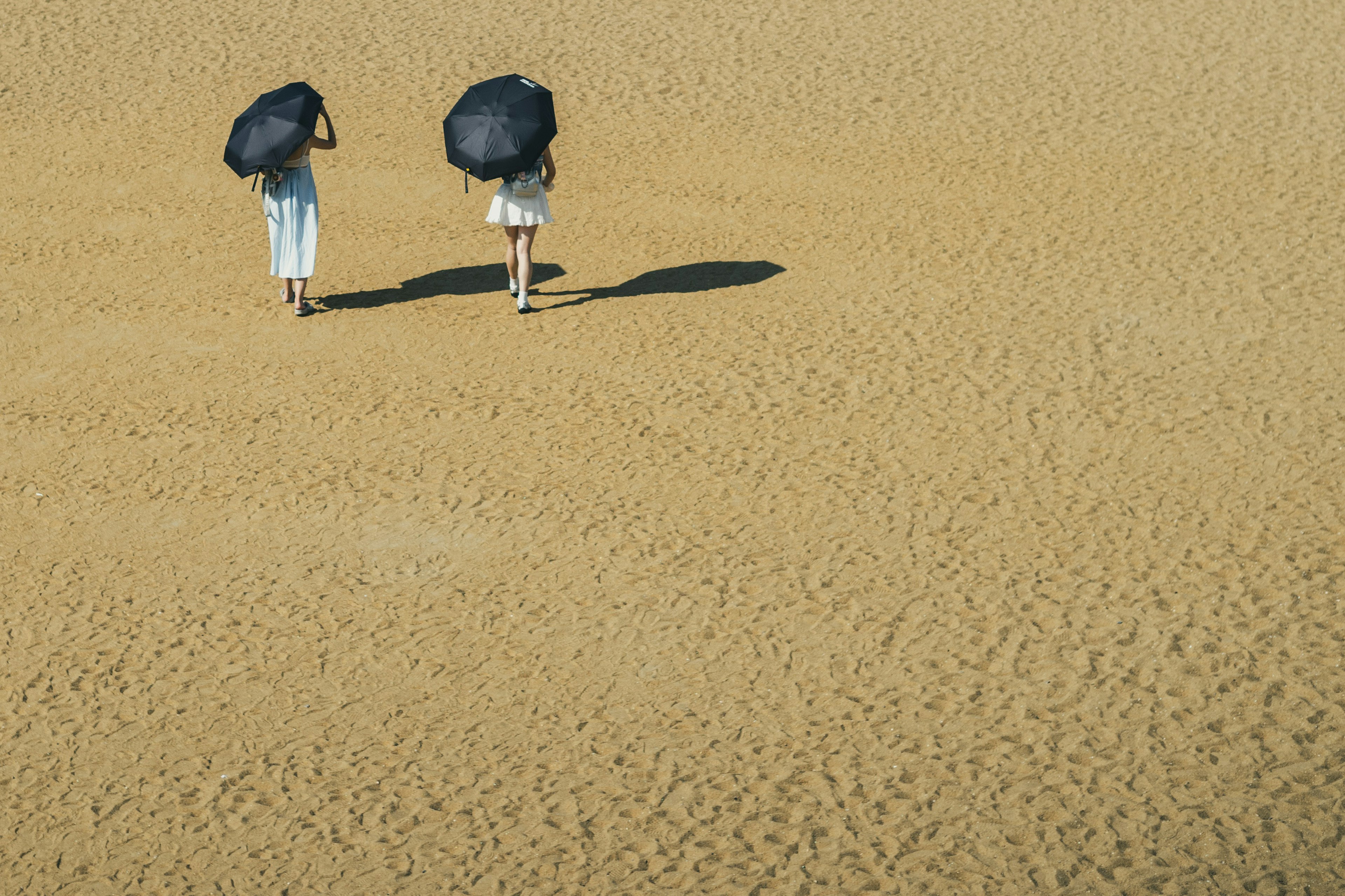 Two women walking on a sandy beach holding black umbrellas