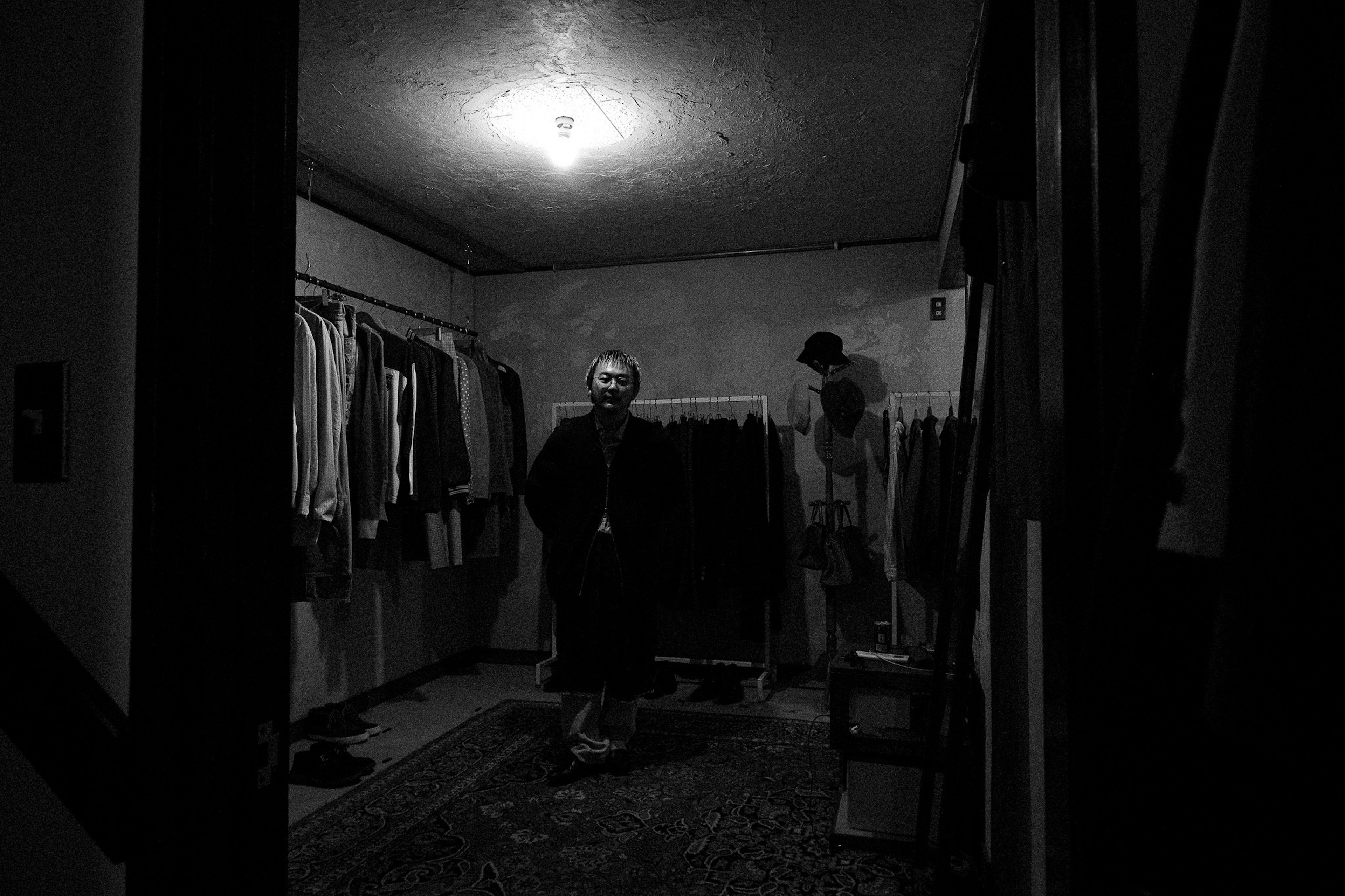 A figure standing in a dimly lit room with clothes hanging in the background