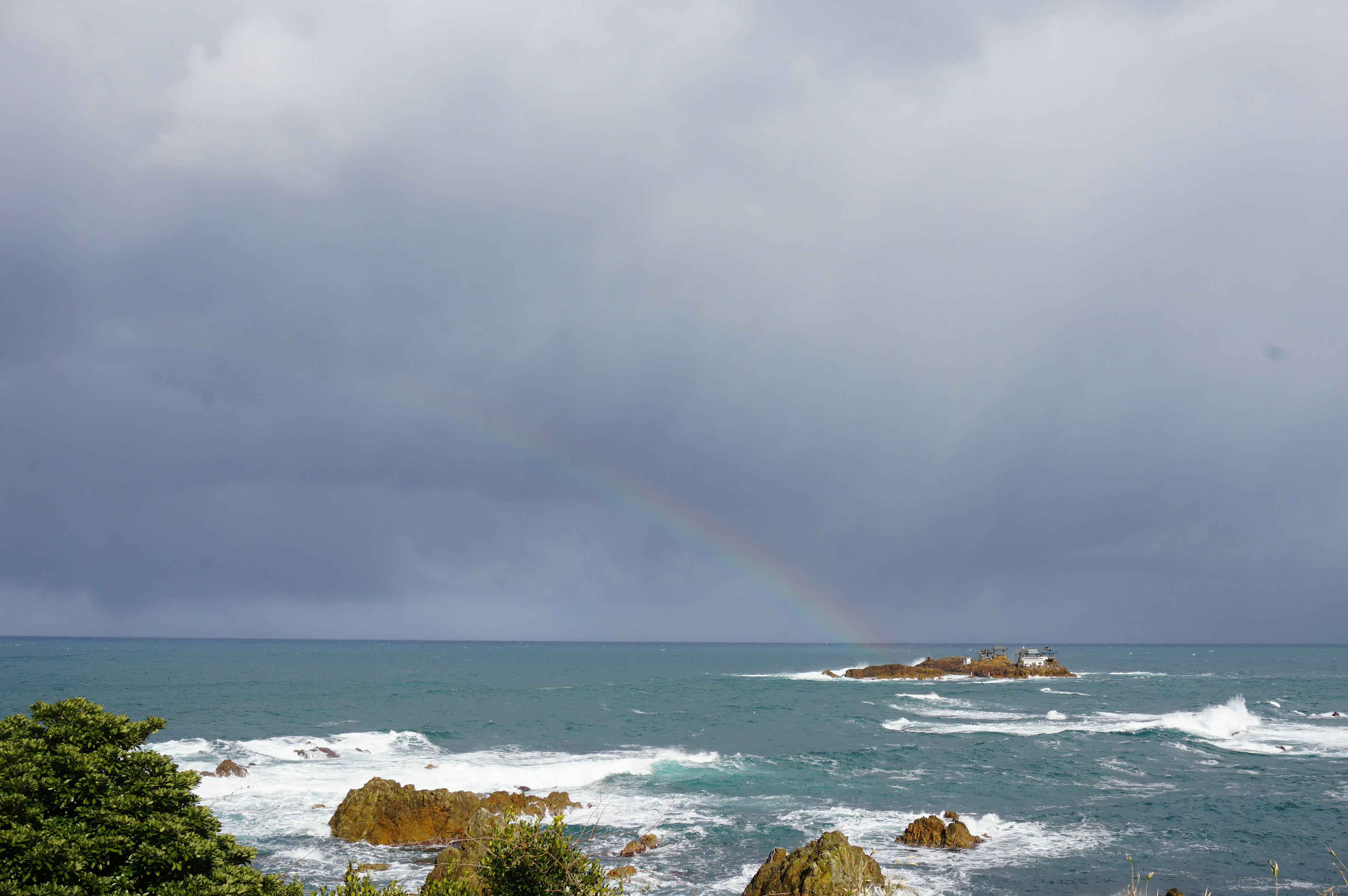 波が立つ海と暗い雲の下に虹がかかる風景