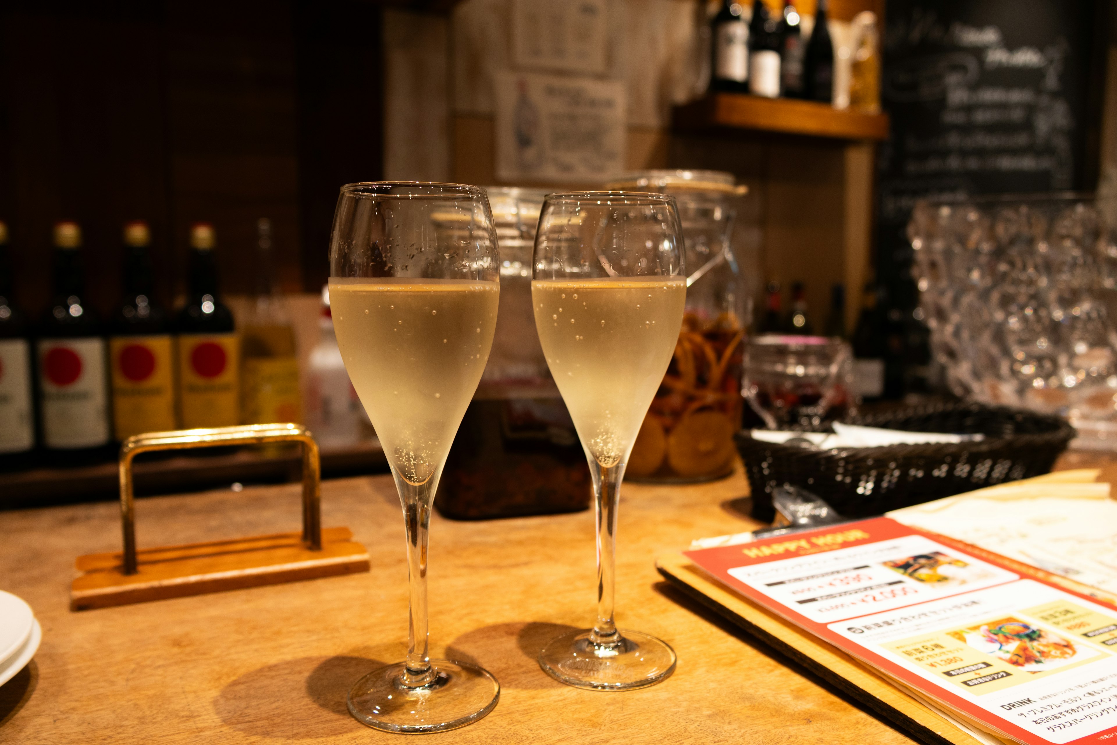 Two champagne glasses on a bar counter with bottles in the background