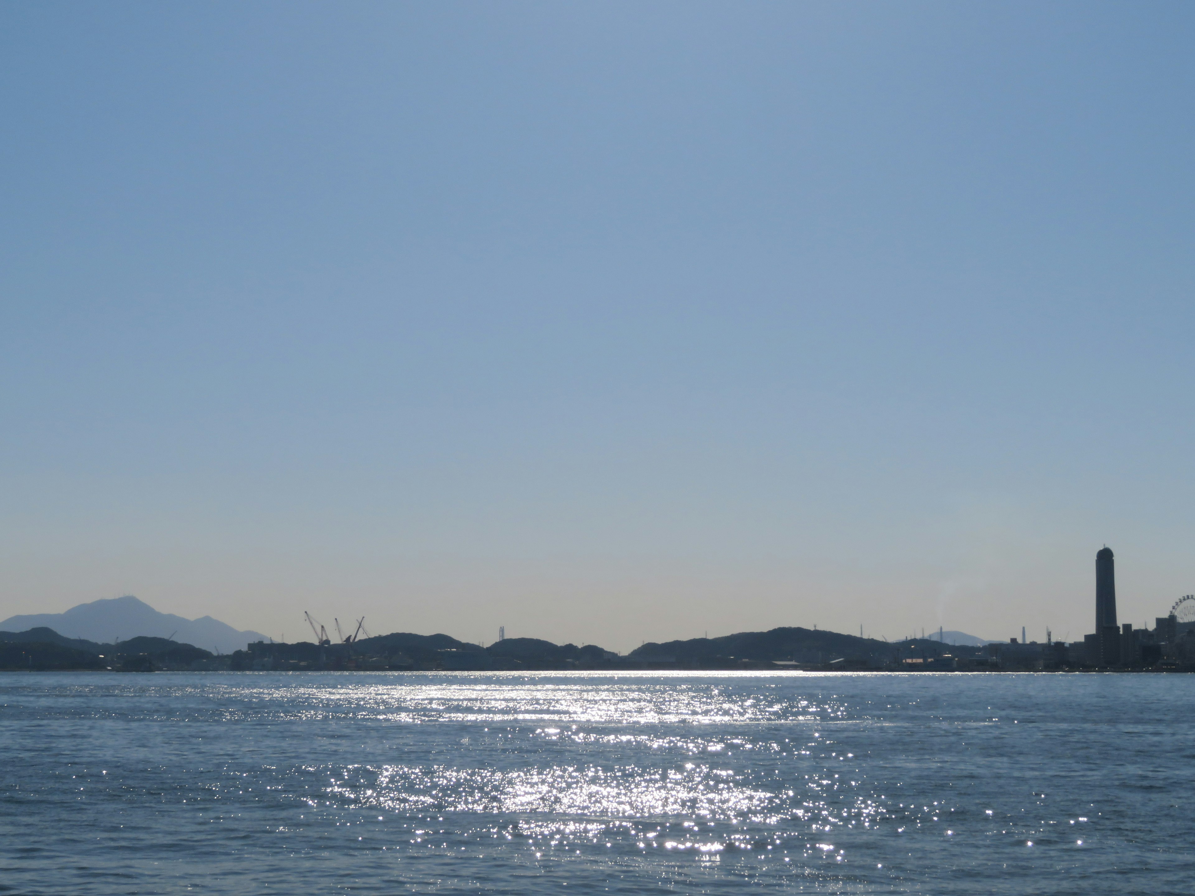 Cielo azul claro sobre agua tranquila con reflejos iluminados por el sol, islas distantes y una chimenea visible