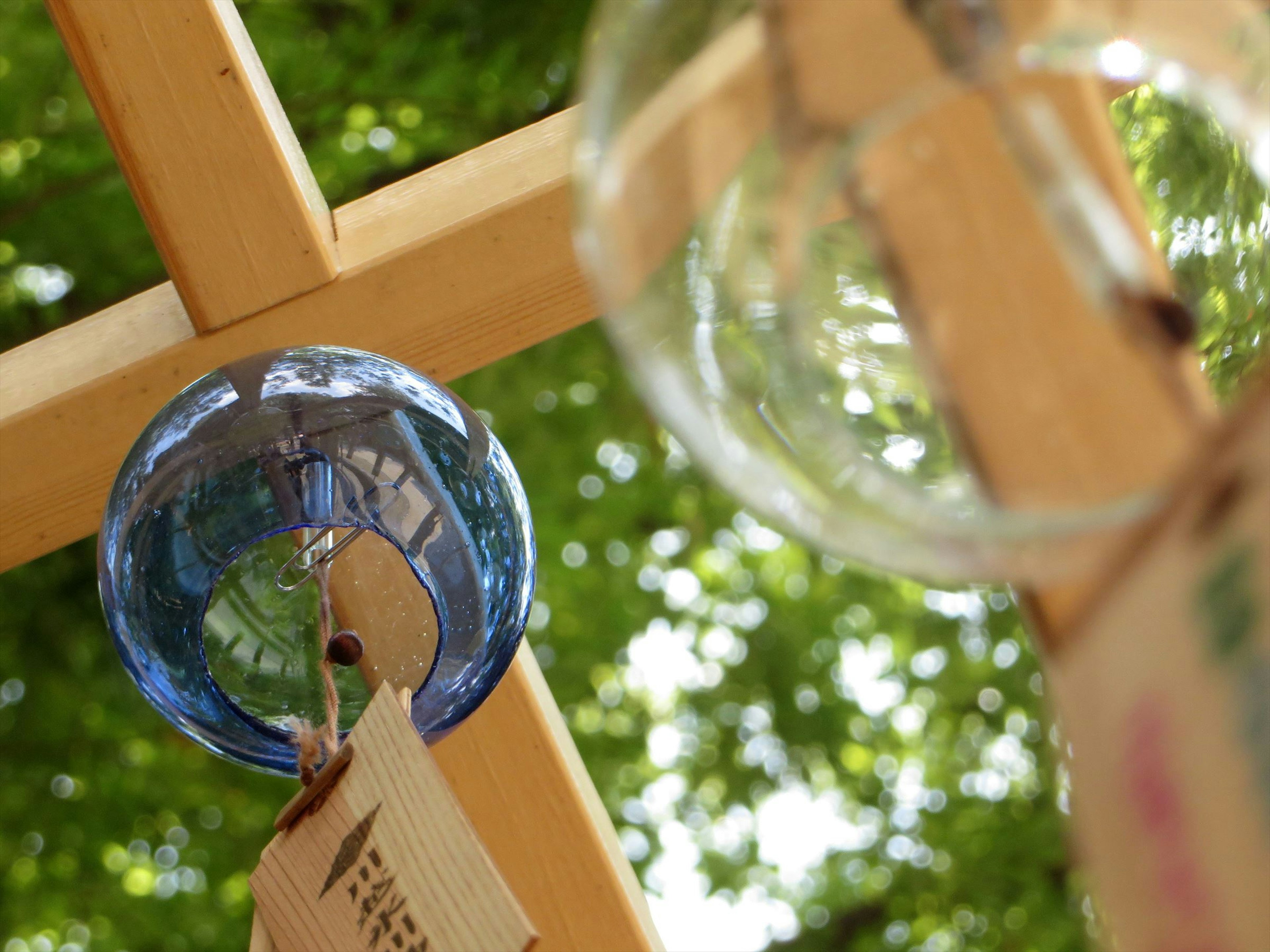 Blaues Glasornament und klares Glasornament hängen von einer Holzstruktur vor grünem Hintergrund