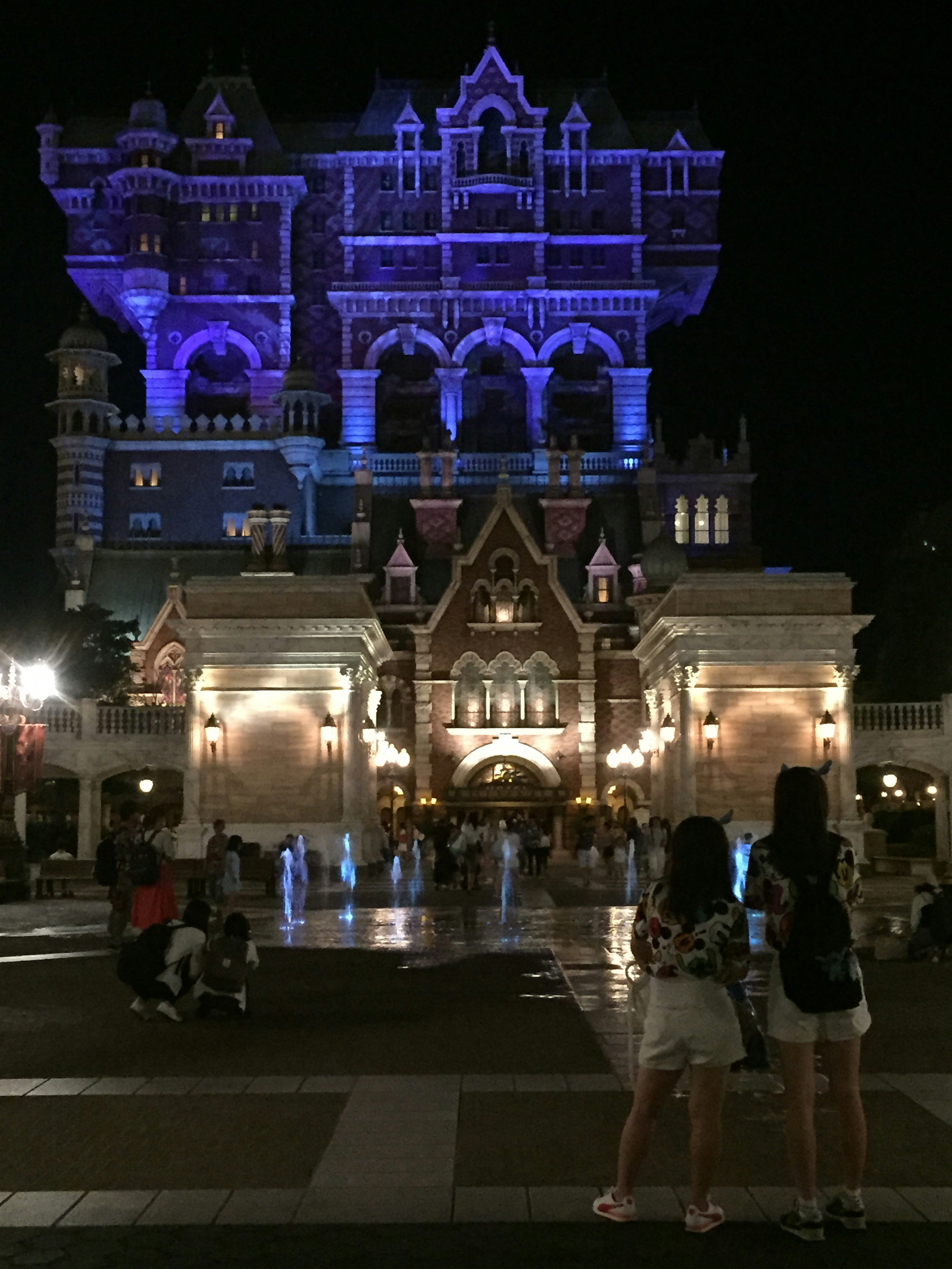 A grand building illuminated in blue and purple at night with two tourists in the foreground