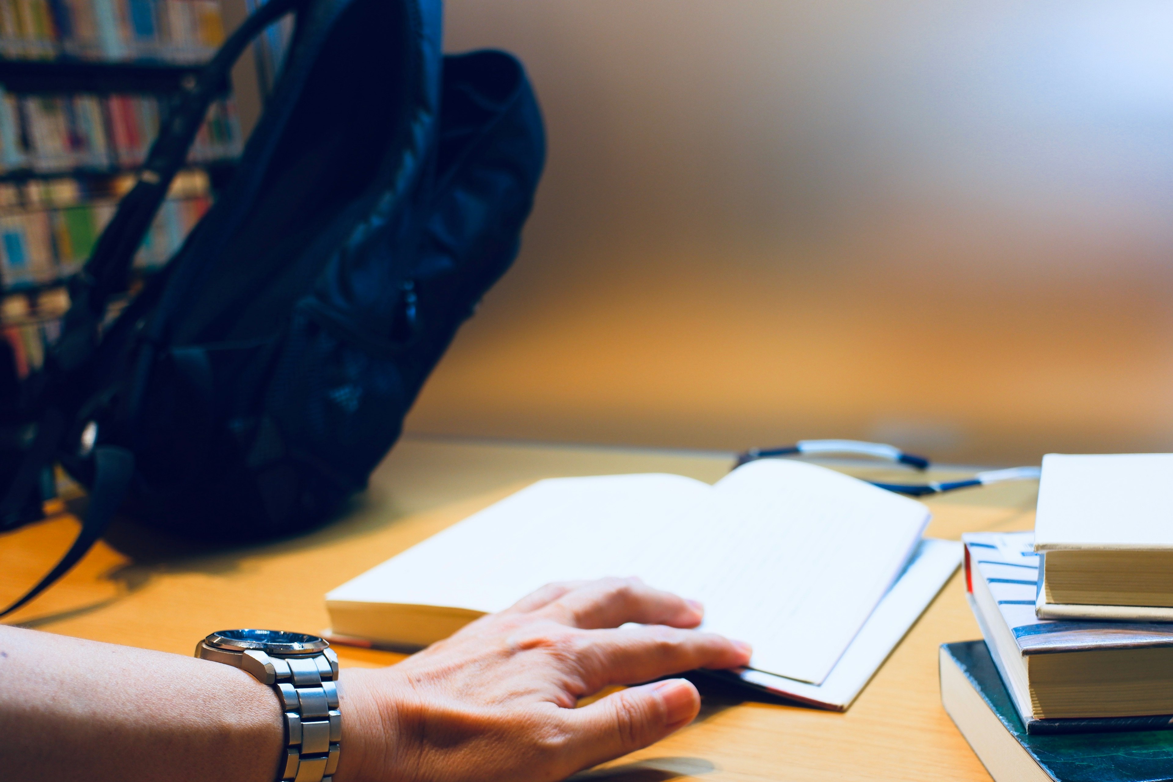 Ein auf dem Tisch liegendes Buch mit einer Hand die sich ihm nähert ein Rucksack in der Nähe und ein Regal im Hintergrund