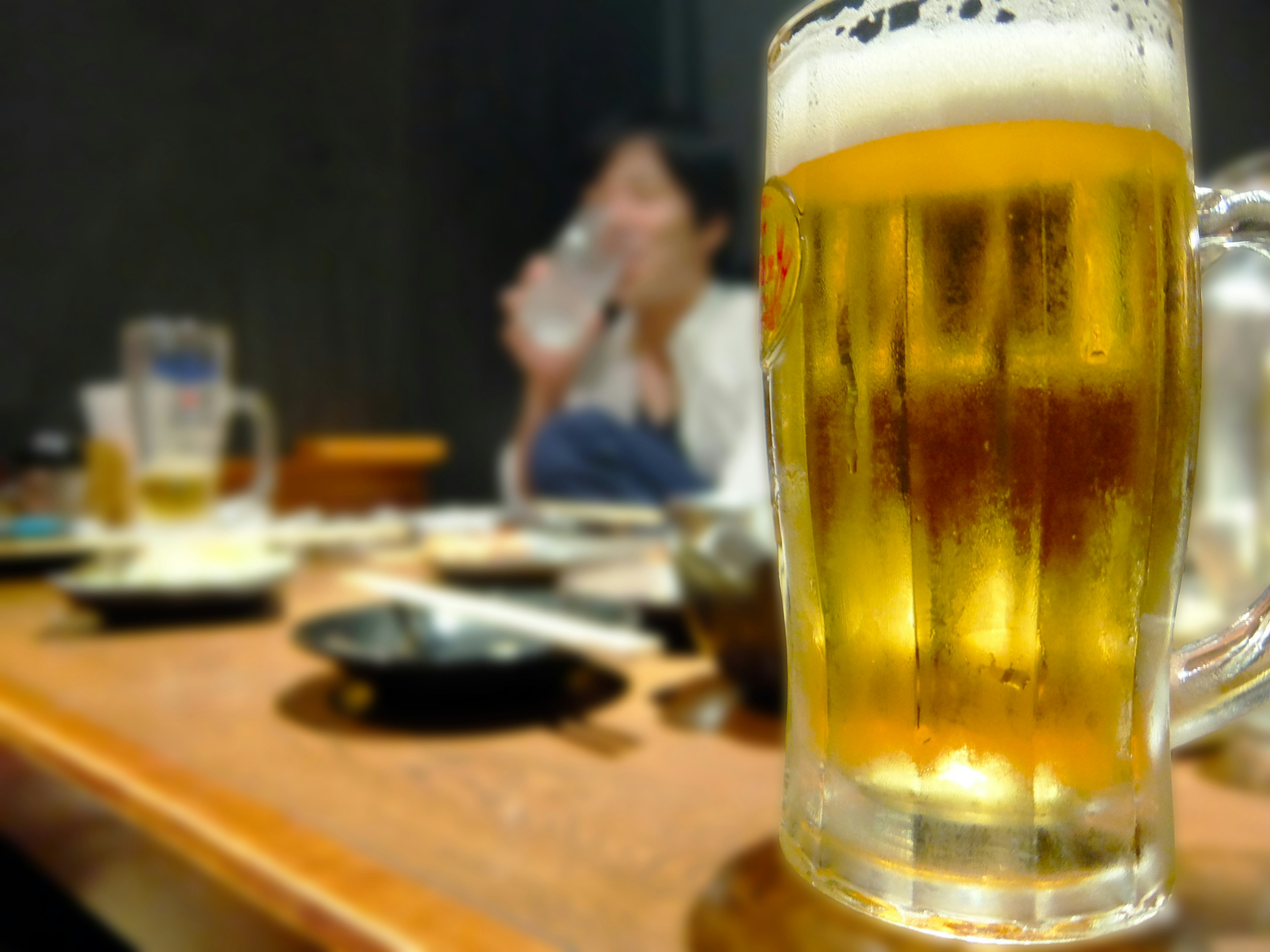 A glass of beer in the foreground with a person drinking water in the background