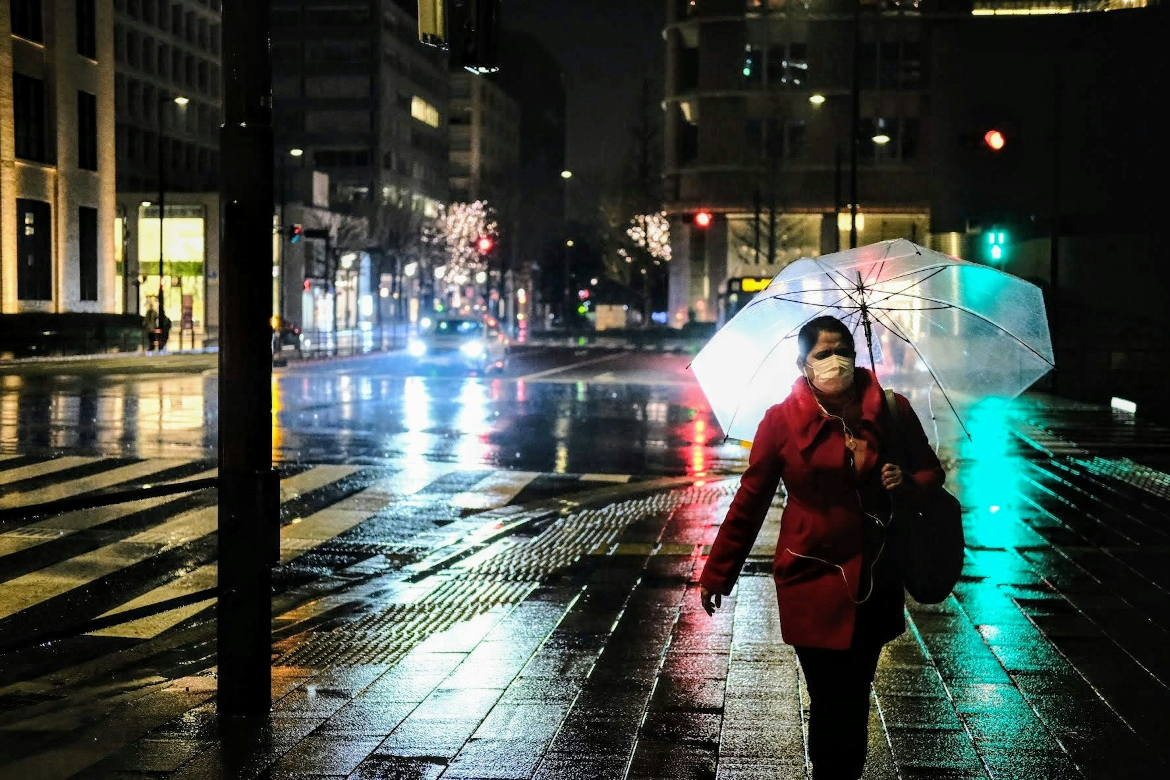 一位穿著紅色外套的女性在夜晚的潮濕城市街道上走，手持透明雨傘