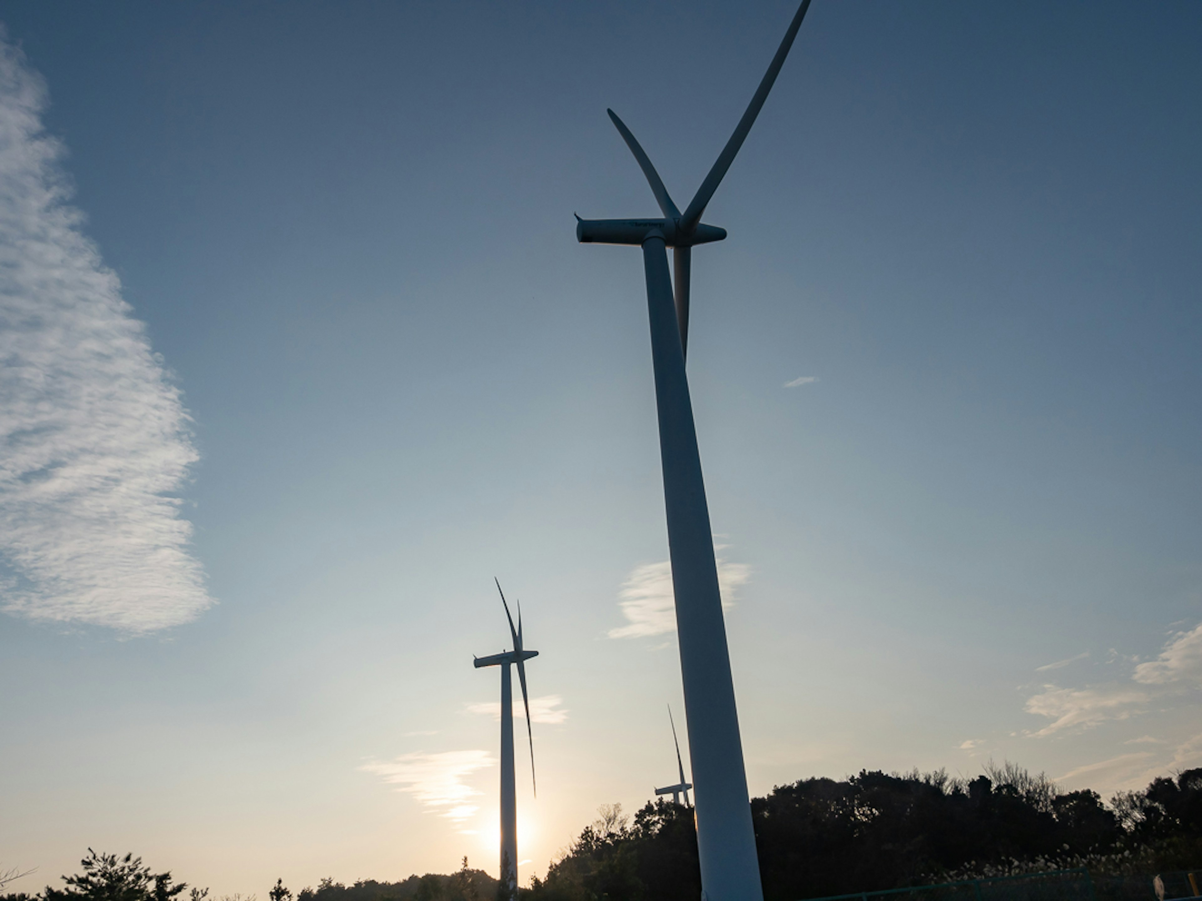 青空の下に立つ風力発電機が二つ見える