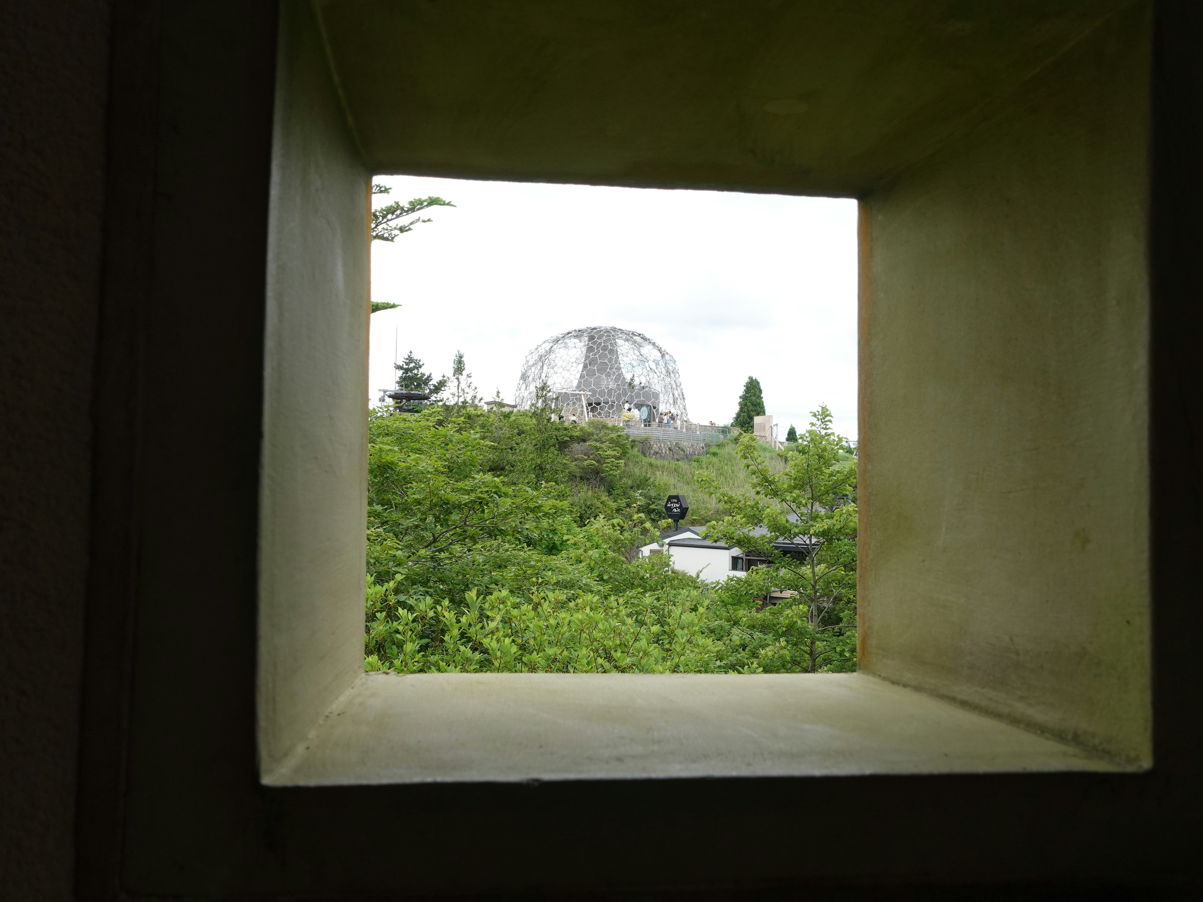 Vista attraverso una finestra quadrata che incornicia una vegetazione lussureggiante e una struttura a forma di cupola