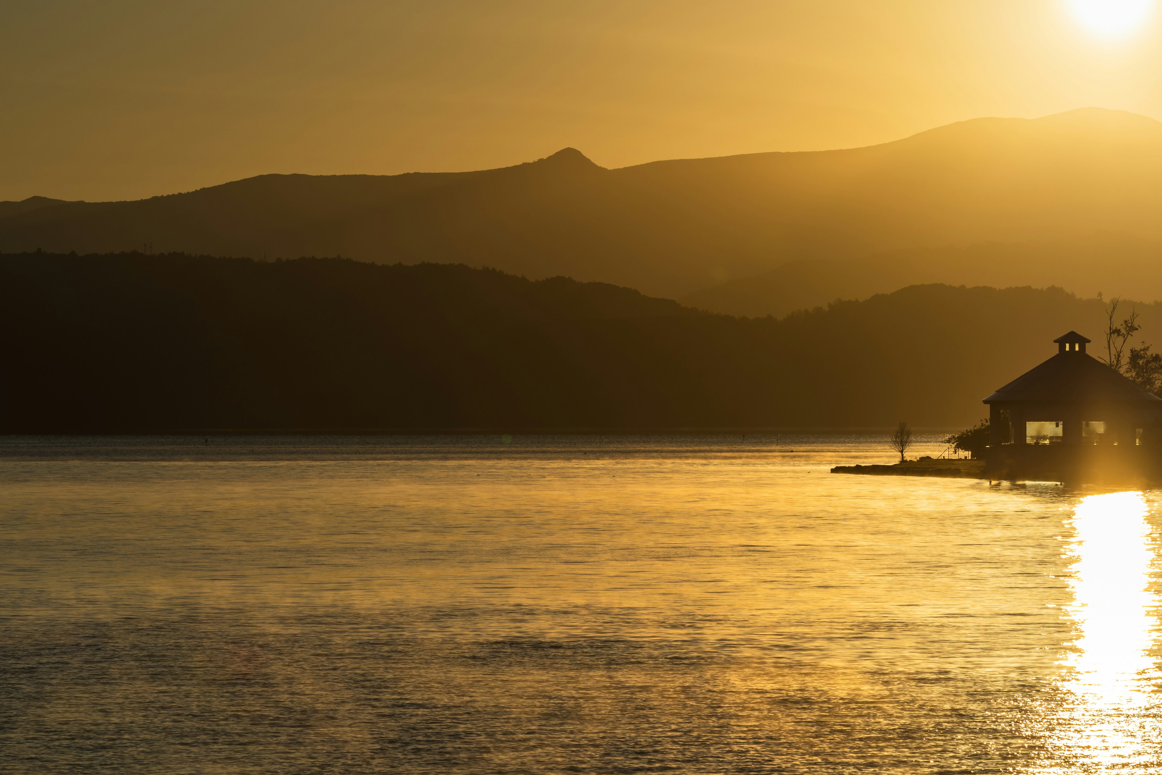 Atardecer sobre un lago tranquilo con montañas y una pequeña cabaña