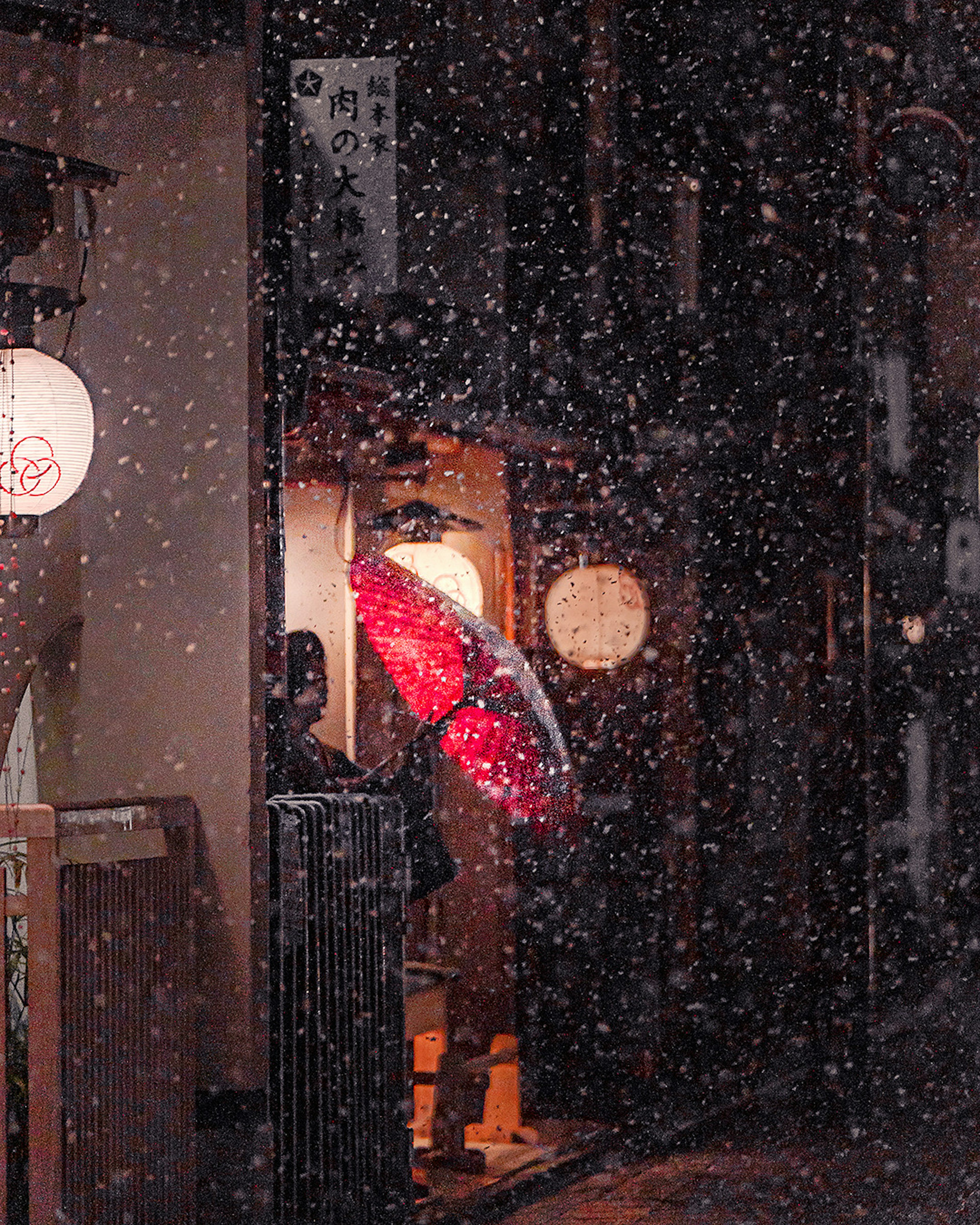 Una persona con un paraguas rojo está de pie en la entrada de una tienda iluminada cálidamente bajo la nieve que cae