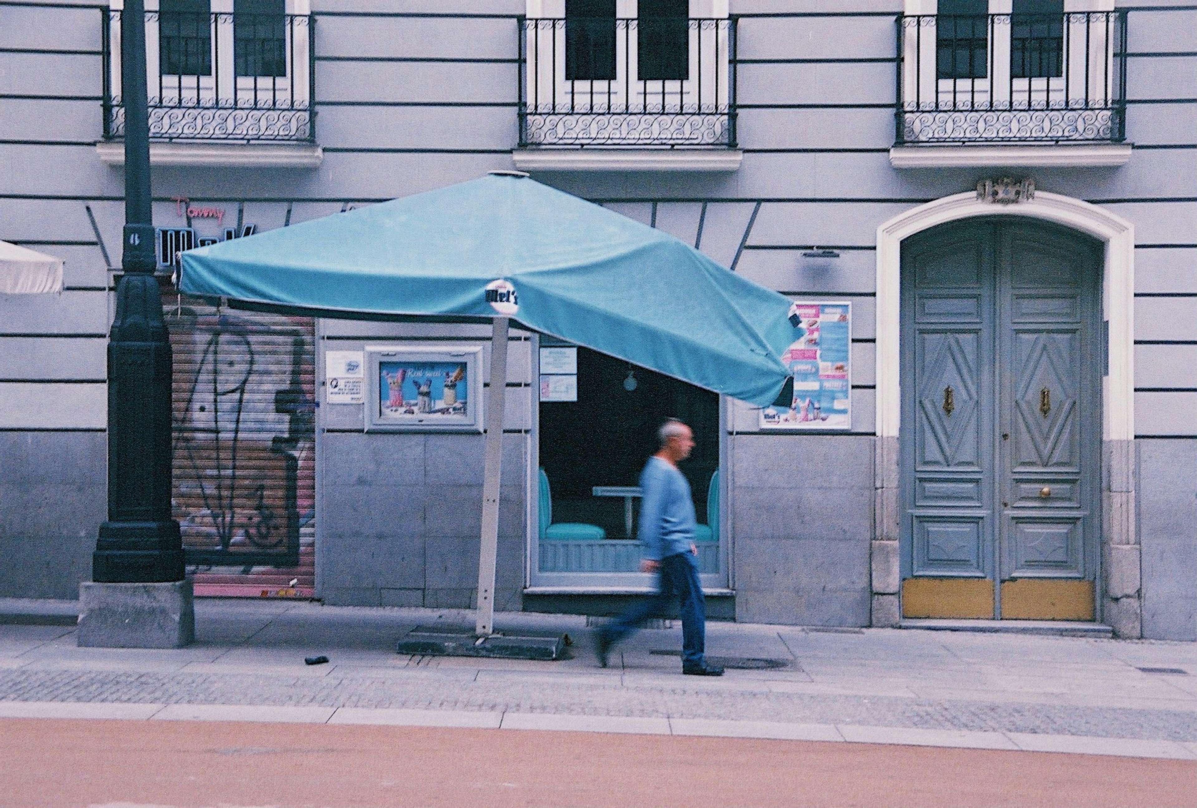 Hombre caminando junto a una tienda azul y la fachada de un edificio gris