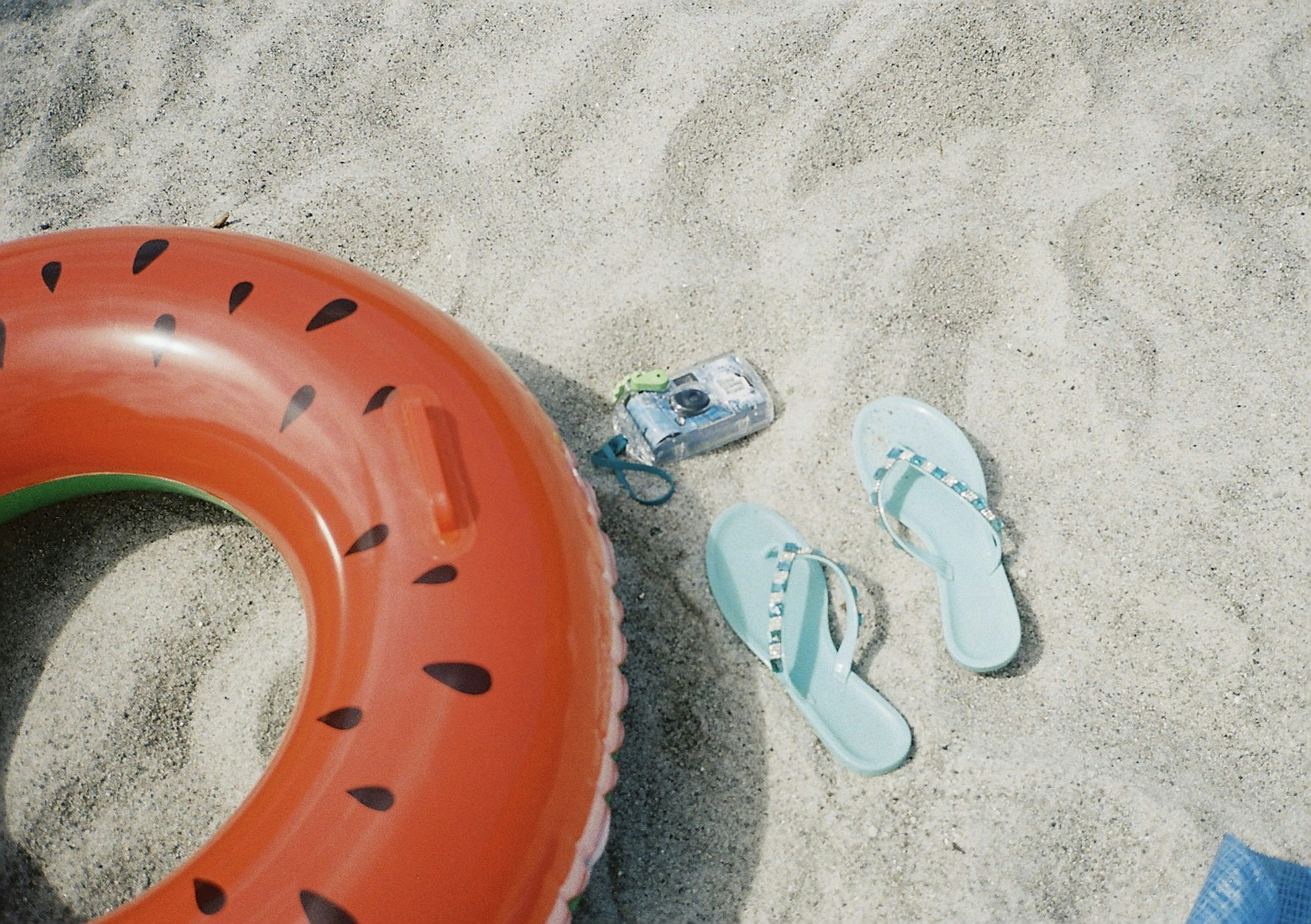 A relaxing beach scene featuring a watermelon float and blue flip-flops on sandy surface