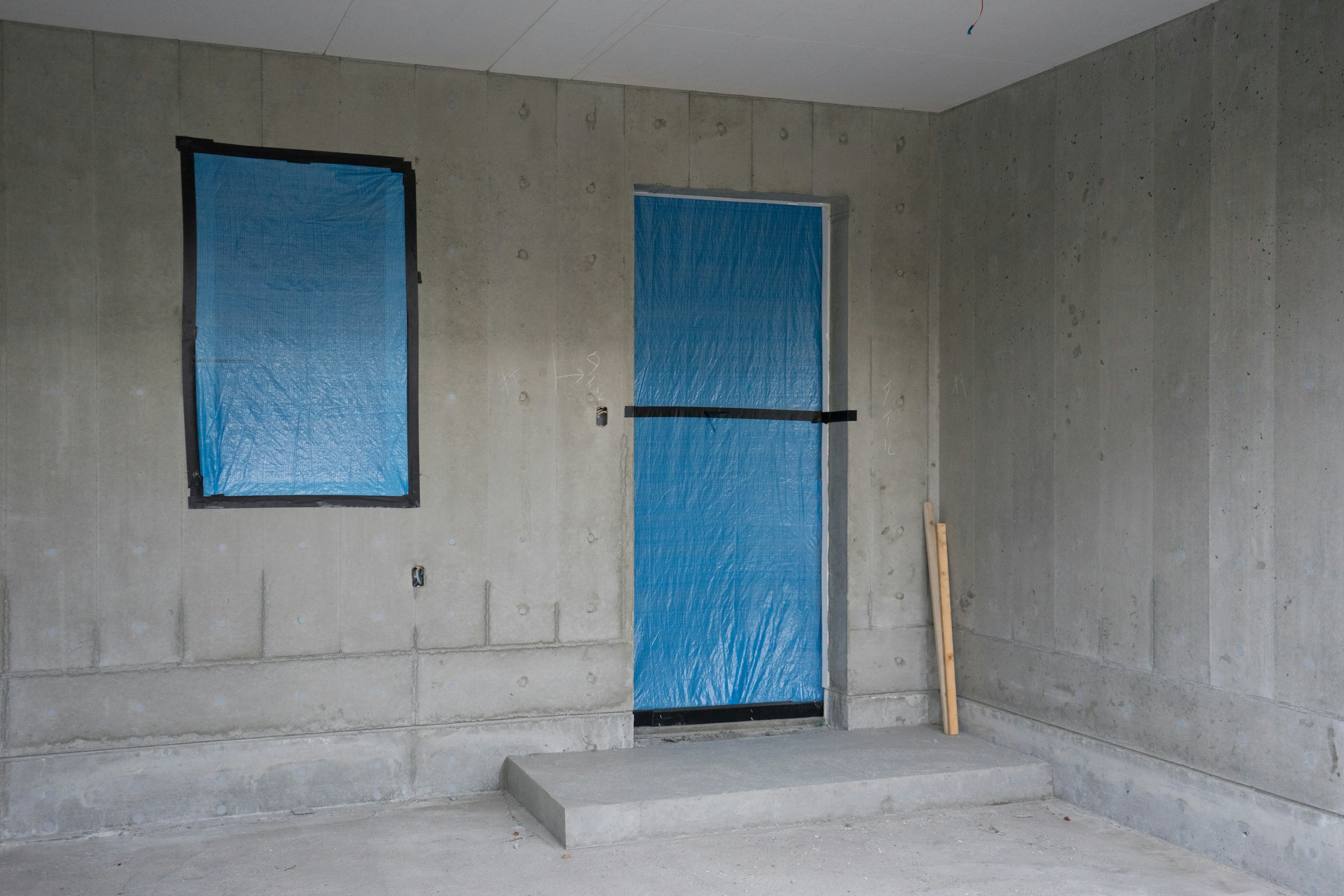 An unfinished room with concrete walls featuring a blue plastic-covered window and door