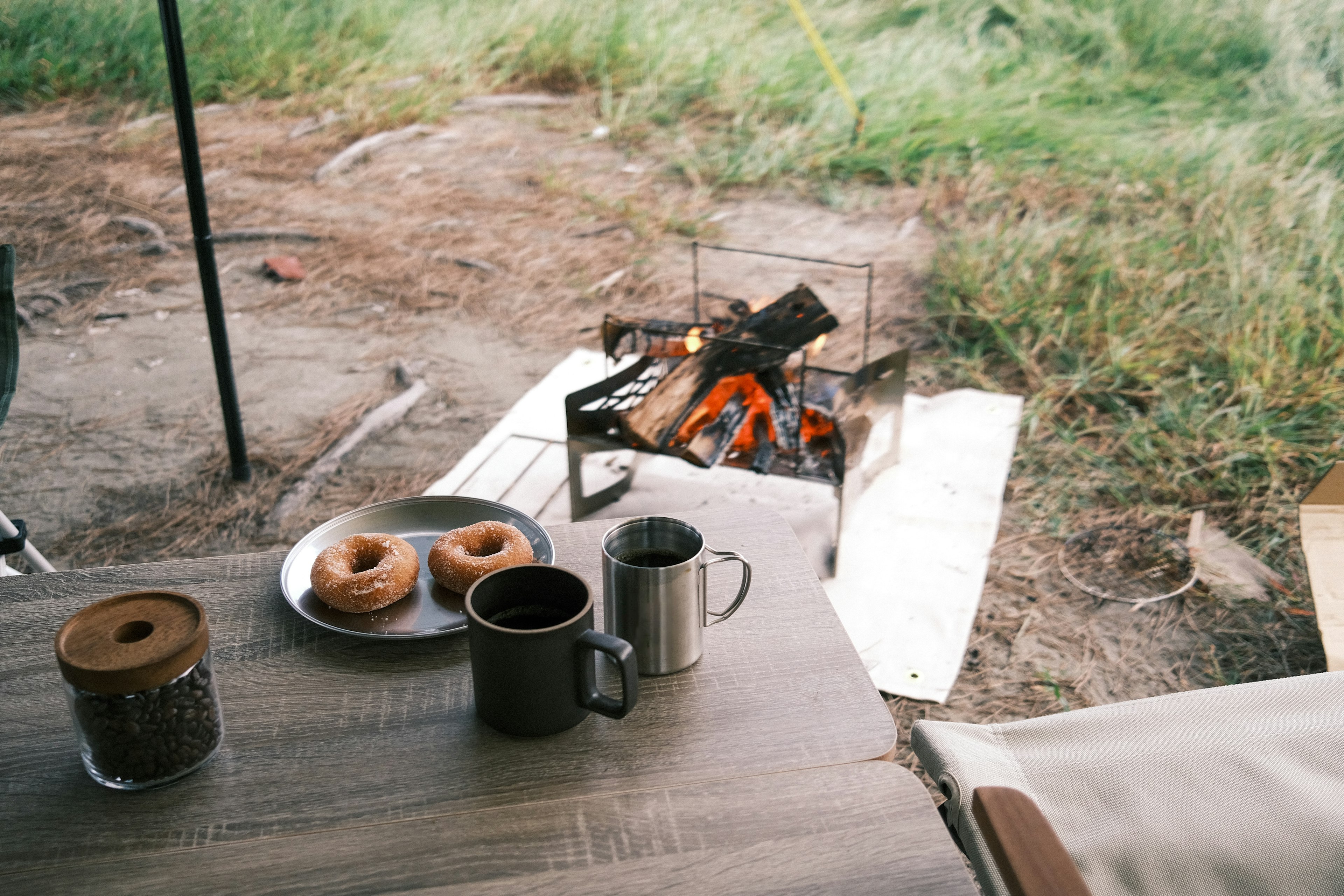 Una mesa de campamento con café y donuts visibles junto a una fogata al fondo