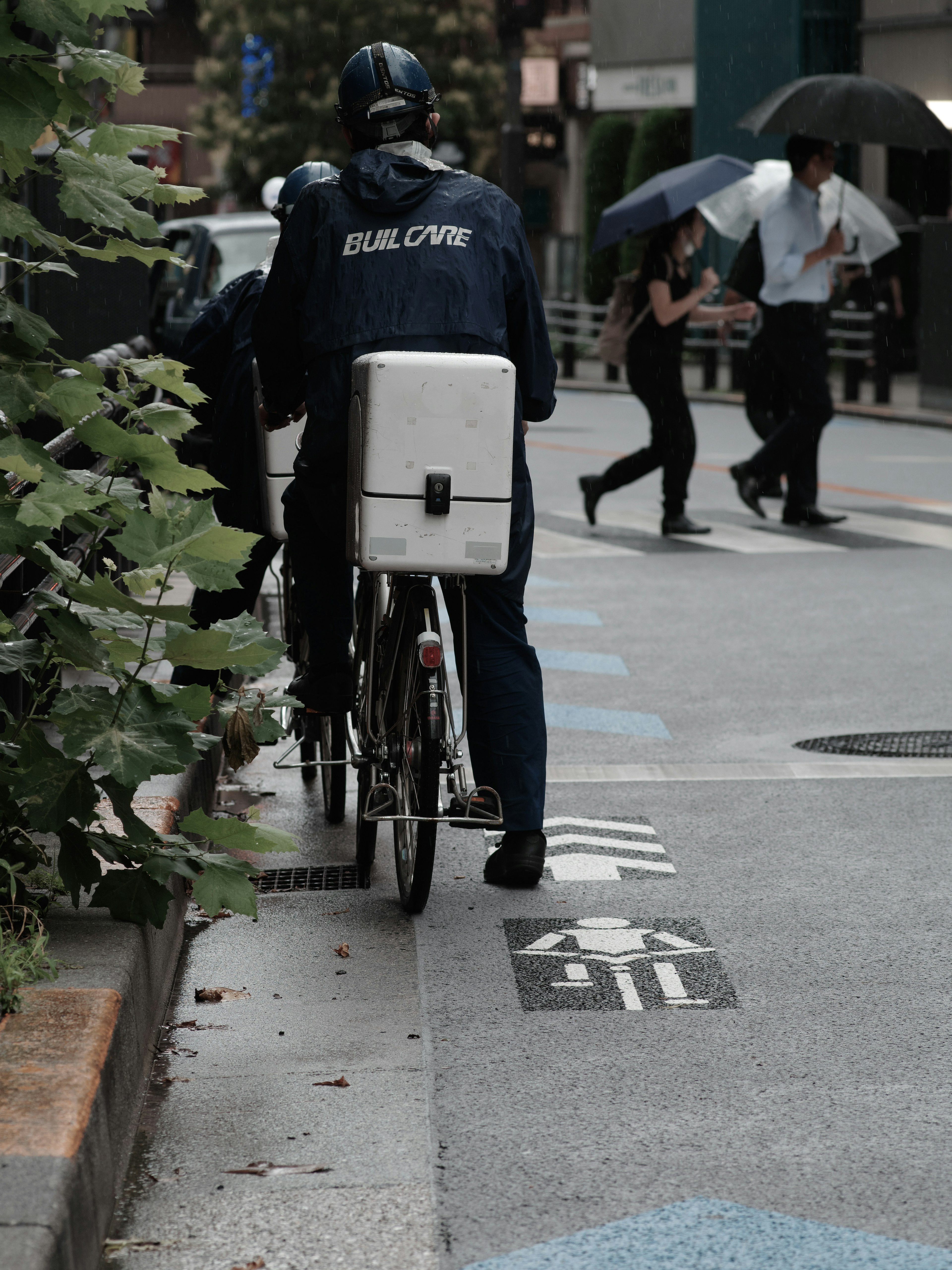 Repartidor en bicicleta con caja de entrega y peatones caminando bajo la lluvia