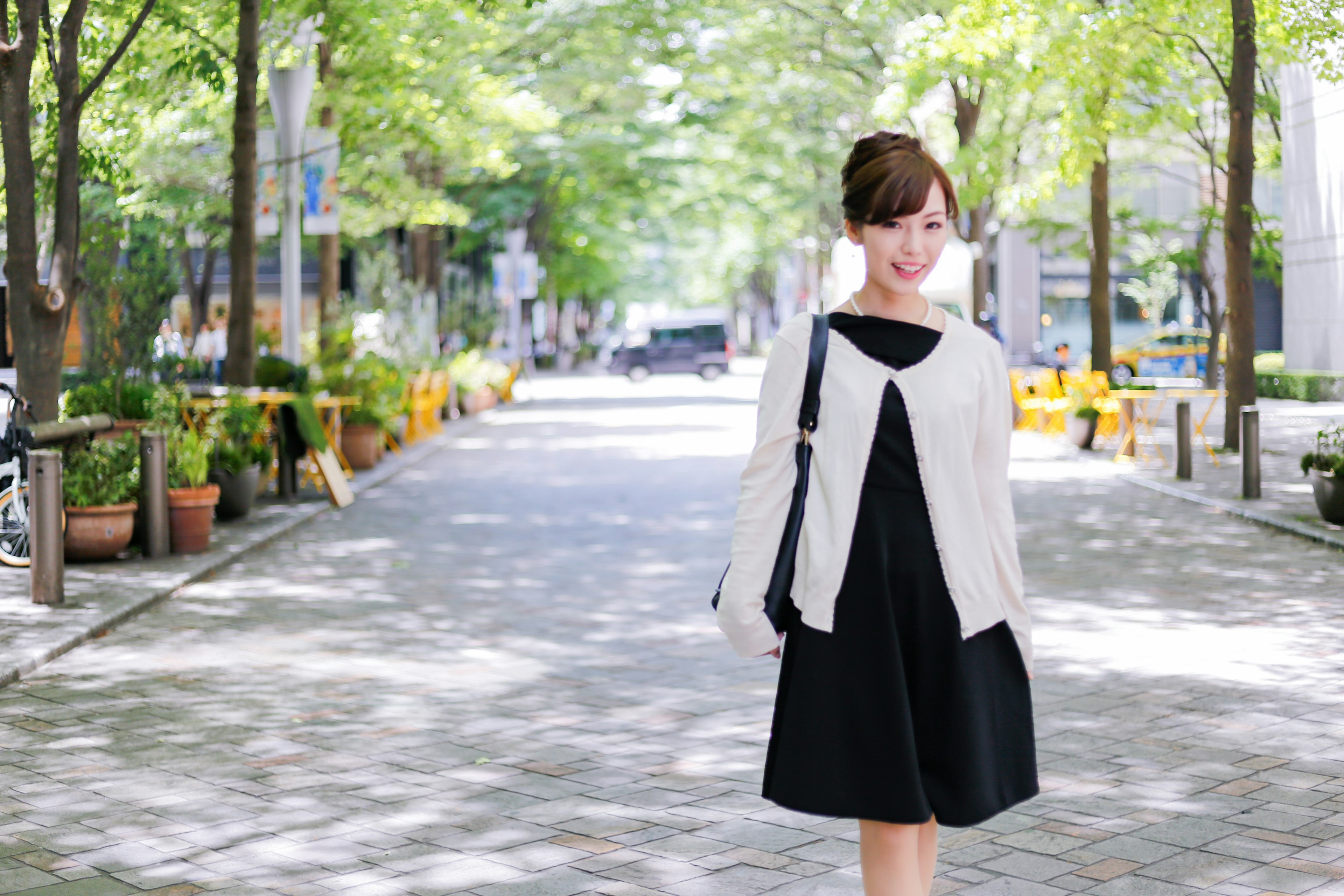 Woman walking along a park path wearing a white cardigan and a black dress surrounded by green trees and potted plants