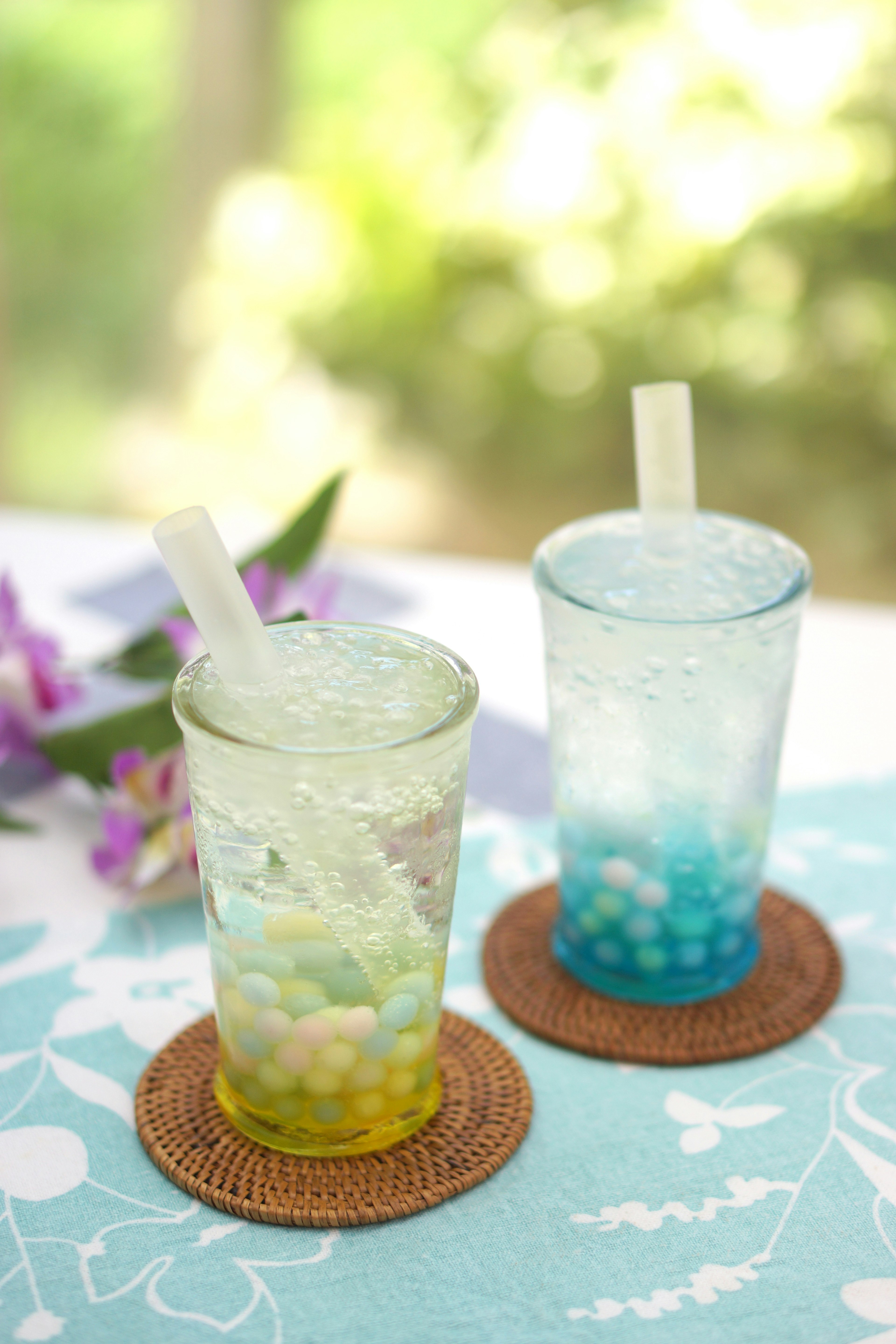 Deux verres avec des boissons colorées aux teintes bleues et vertes placés sur des dessous de verre en osier