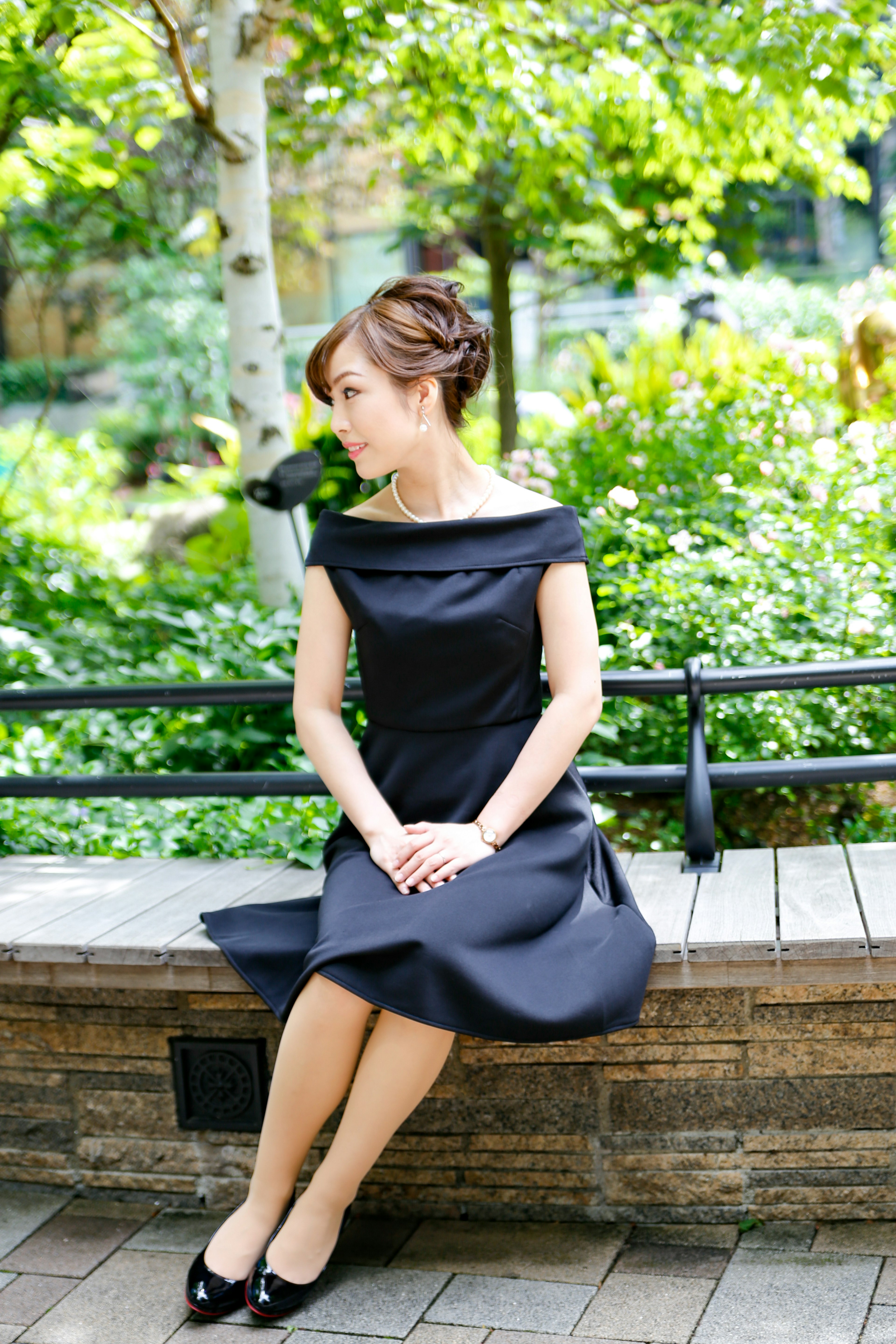 Femme en robe noire assise sur un banc de parc entourée de verdure