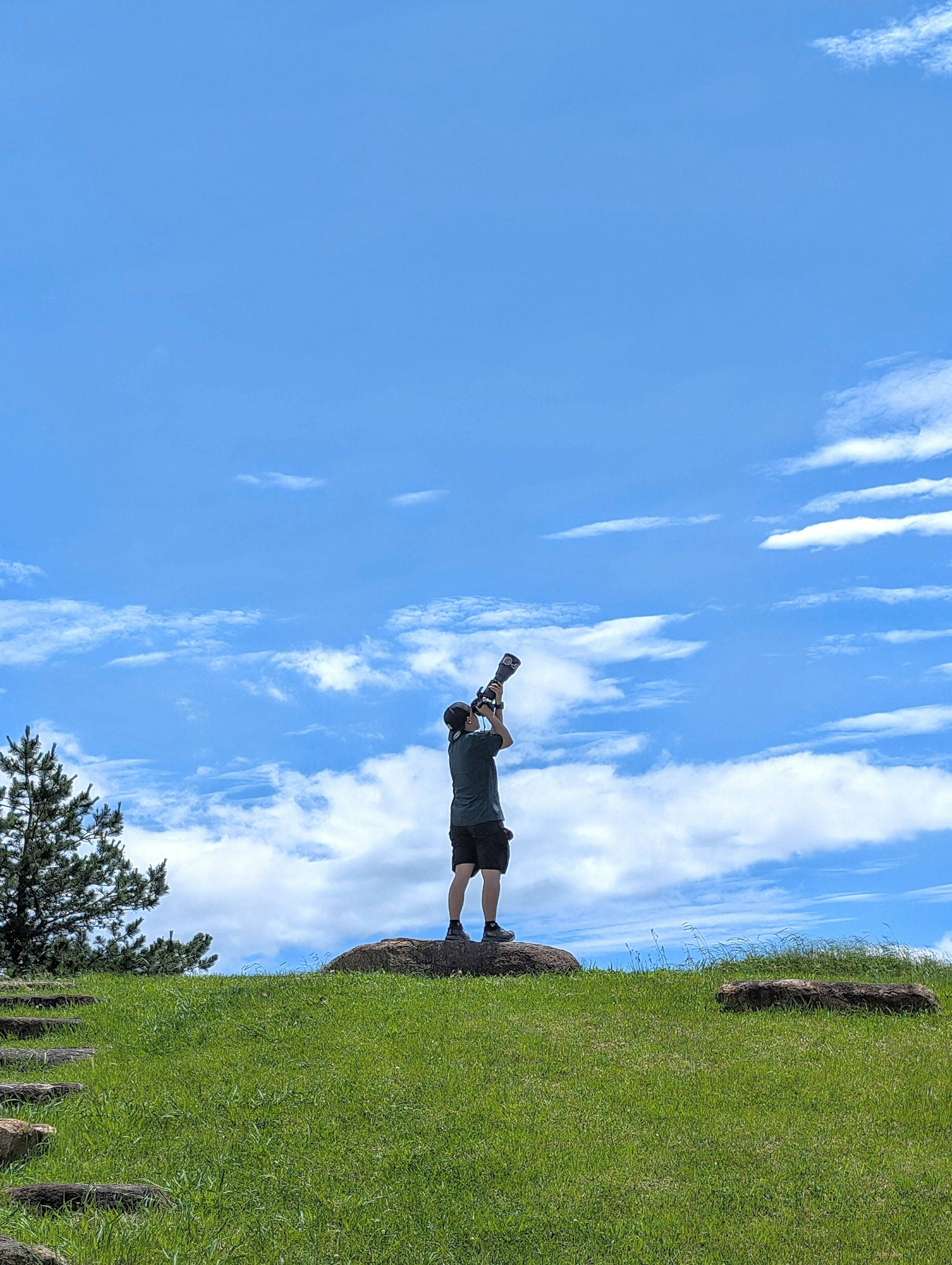 Ein Mann steht auf einem grasbewachsenen Hügel unter einem blauen Himmel und hält eine Kamera