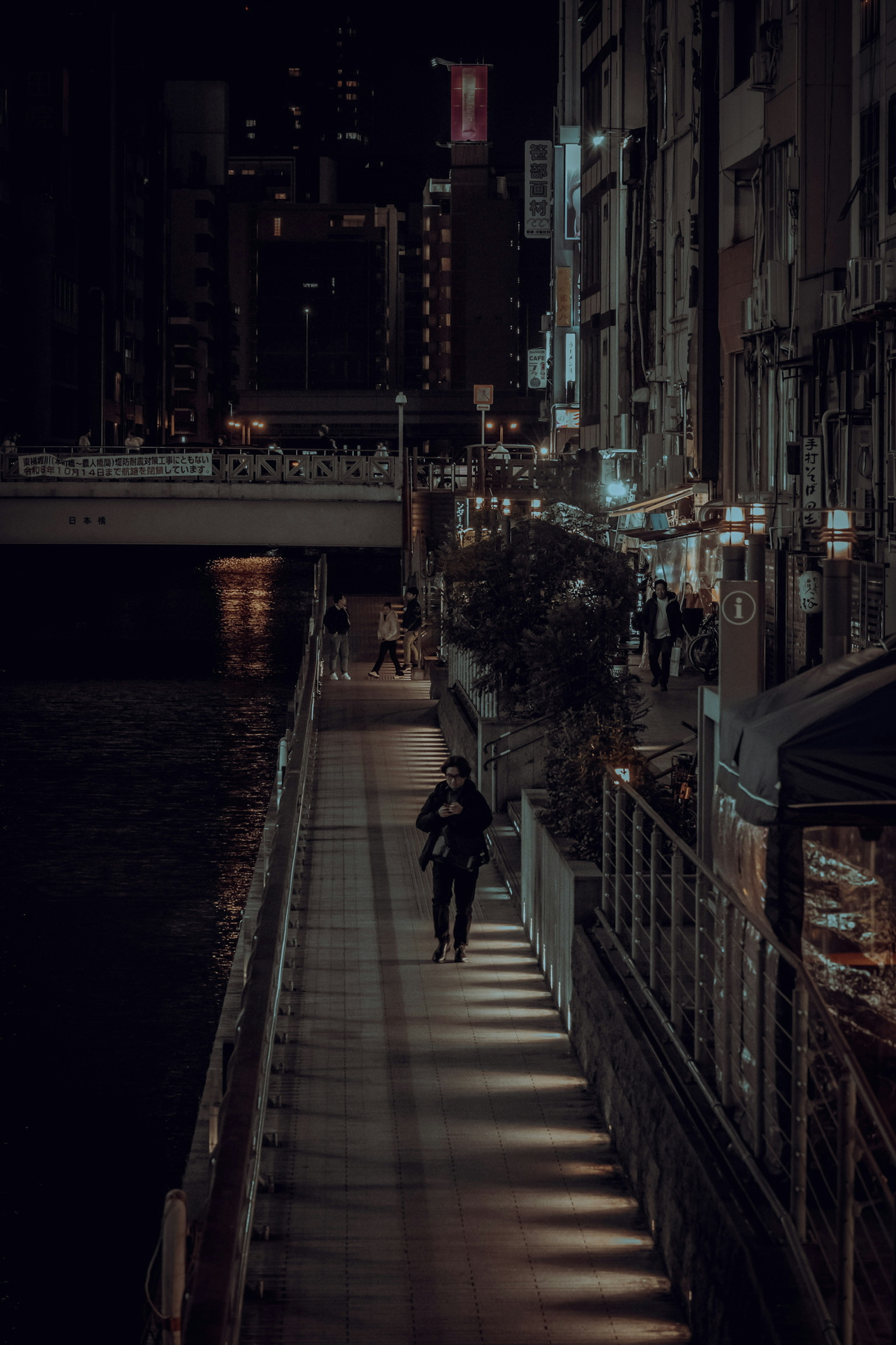 夜の川沿いの歩道を歩く人と明るい街灯