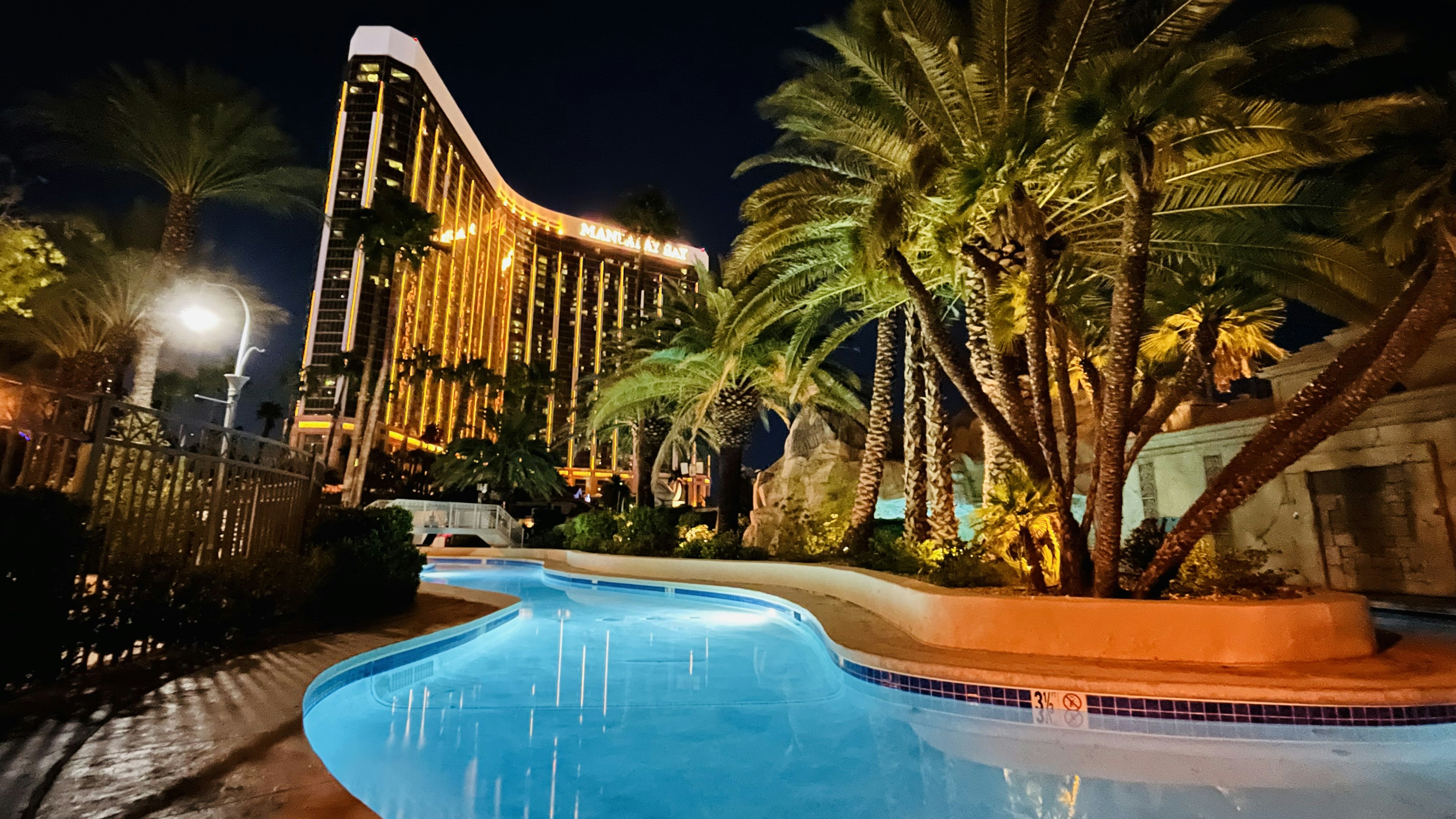 Vista nocturna de una piscina con un hotel alto iluminado y palmeras