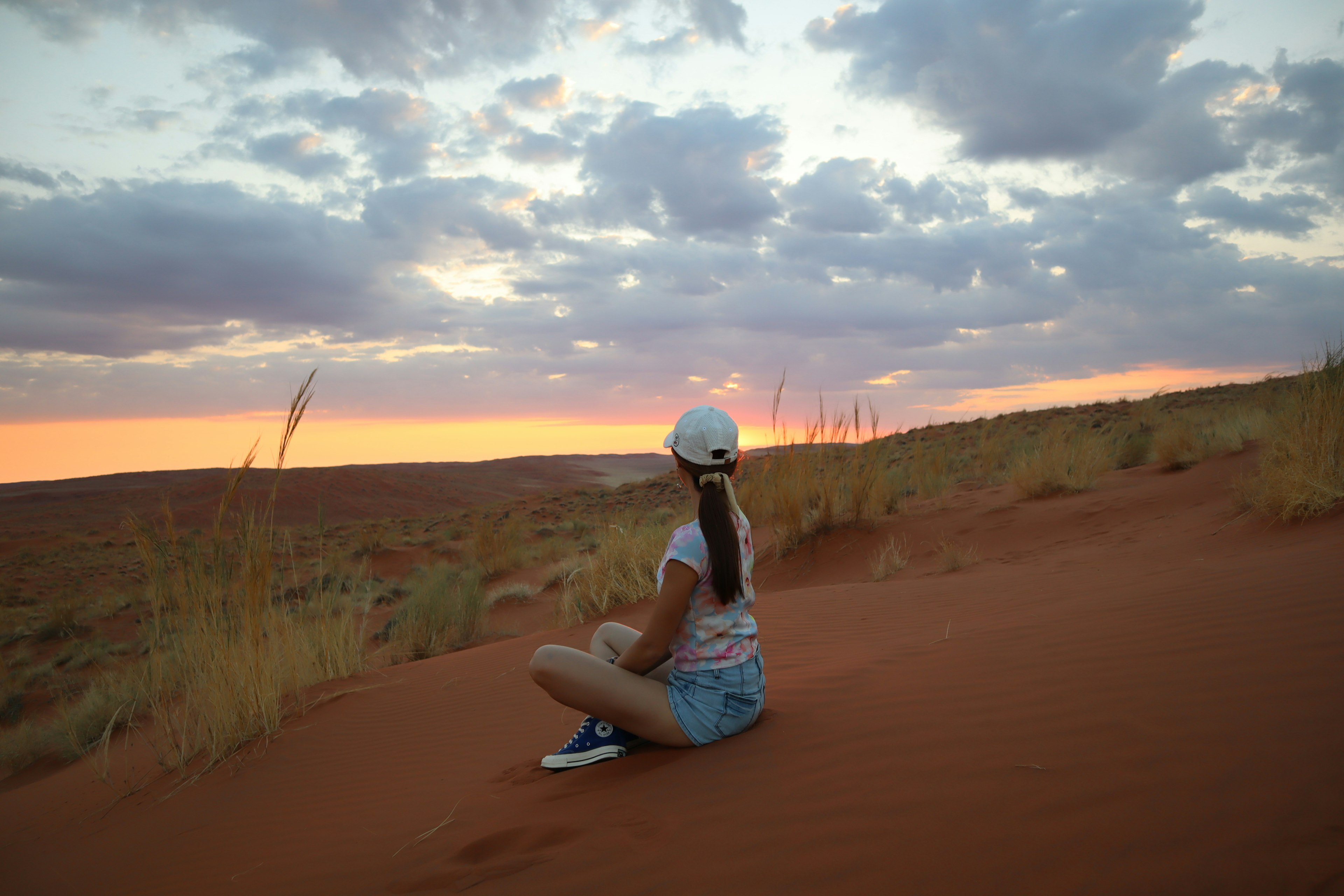 Mädchen sitzt im roten Sand und schaut den Sonnenuntergang an Entspannt in der Natur mit Gras und Dünen