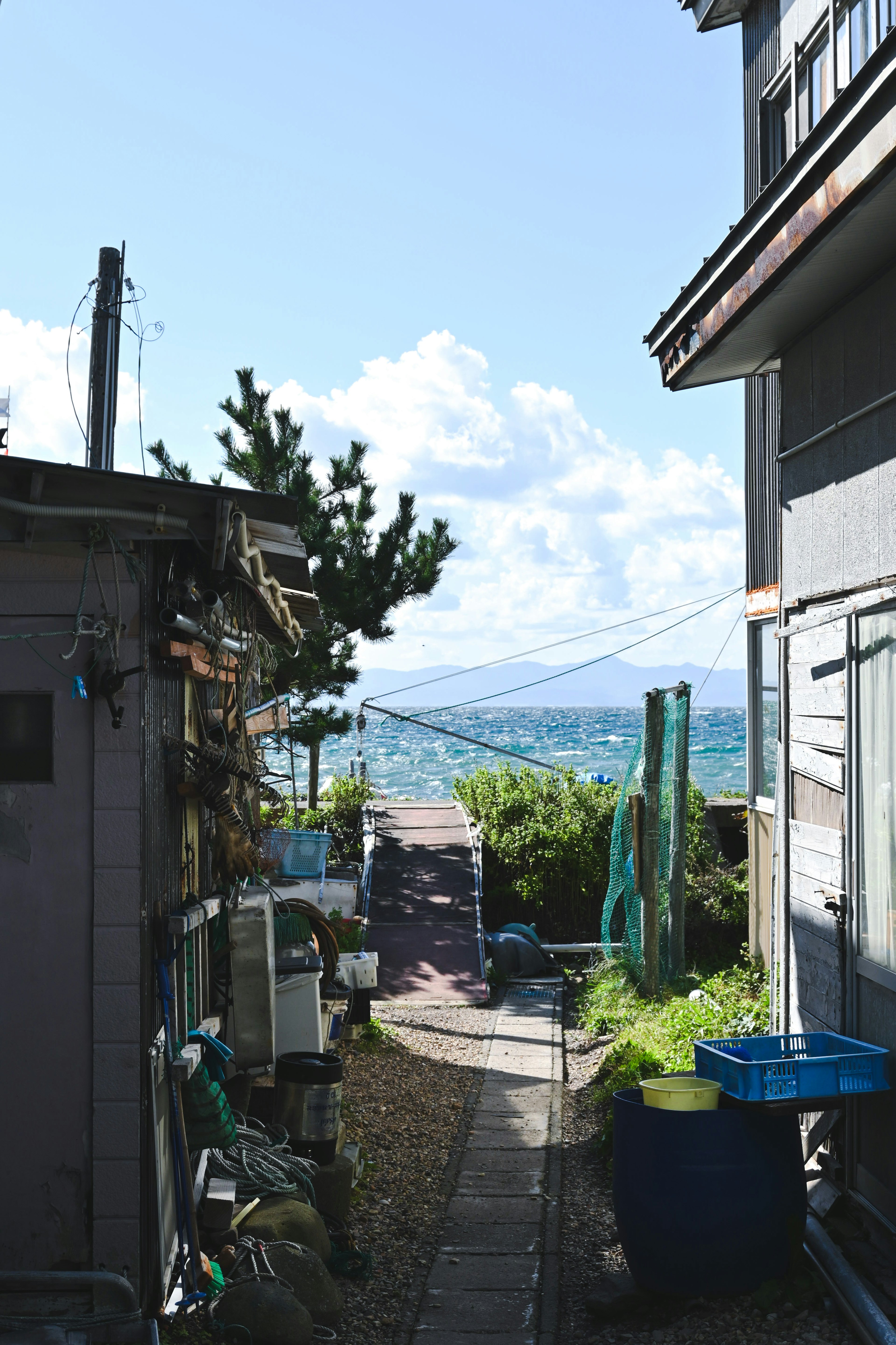 Sentier étroit menant à la mer sous un ciel bleu, bâtiment ancien à gauche, bâtiment moderne à droite