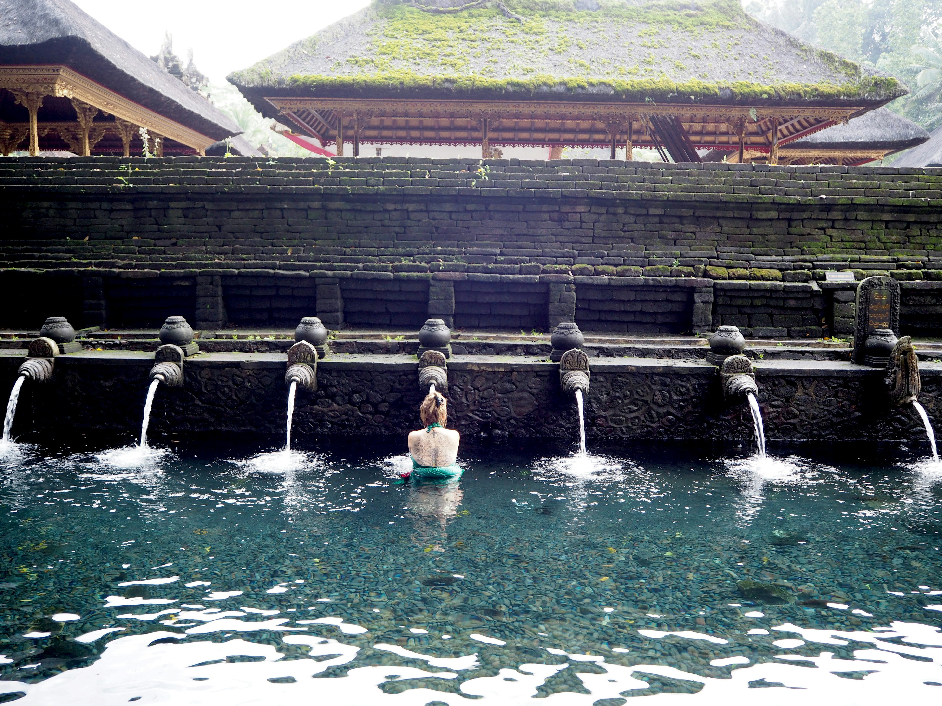 Eine Frau sitzt in einer ruhigen Wasserquelle mit alten Gebäuden im Hintergrund ruhiges und klares Wasser