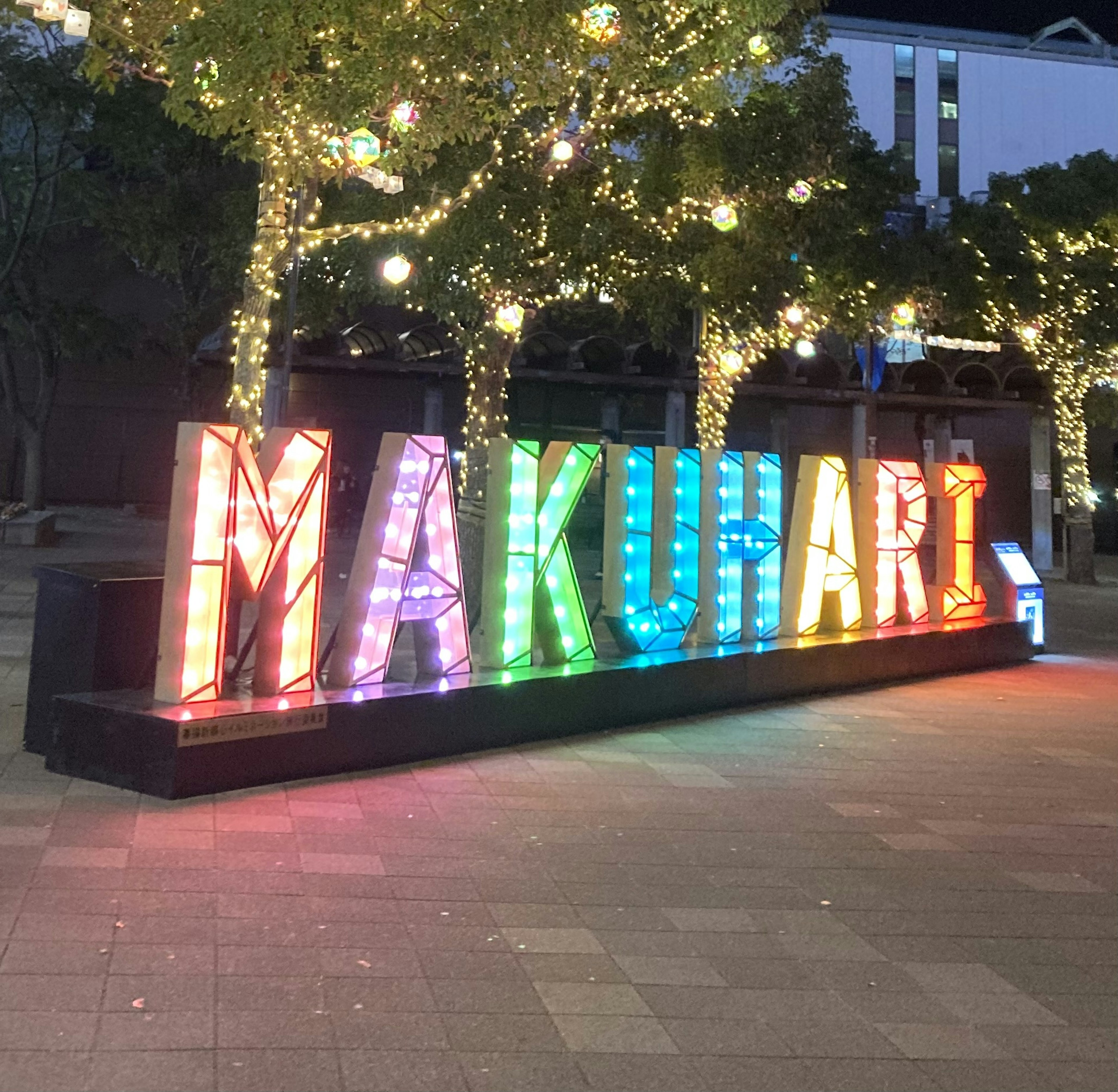 ป้าย 'MAKUHARI' ที่มีสีสันสว่างไสวในเวลากลางคืนพร้อมต้นไม้ที่ประดับ