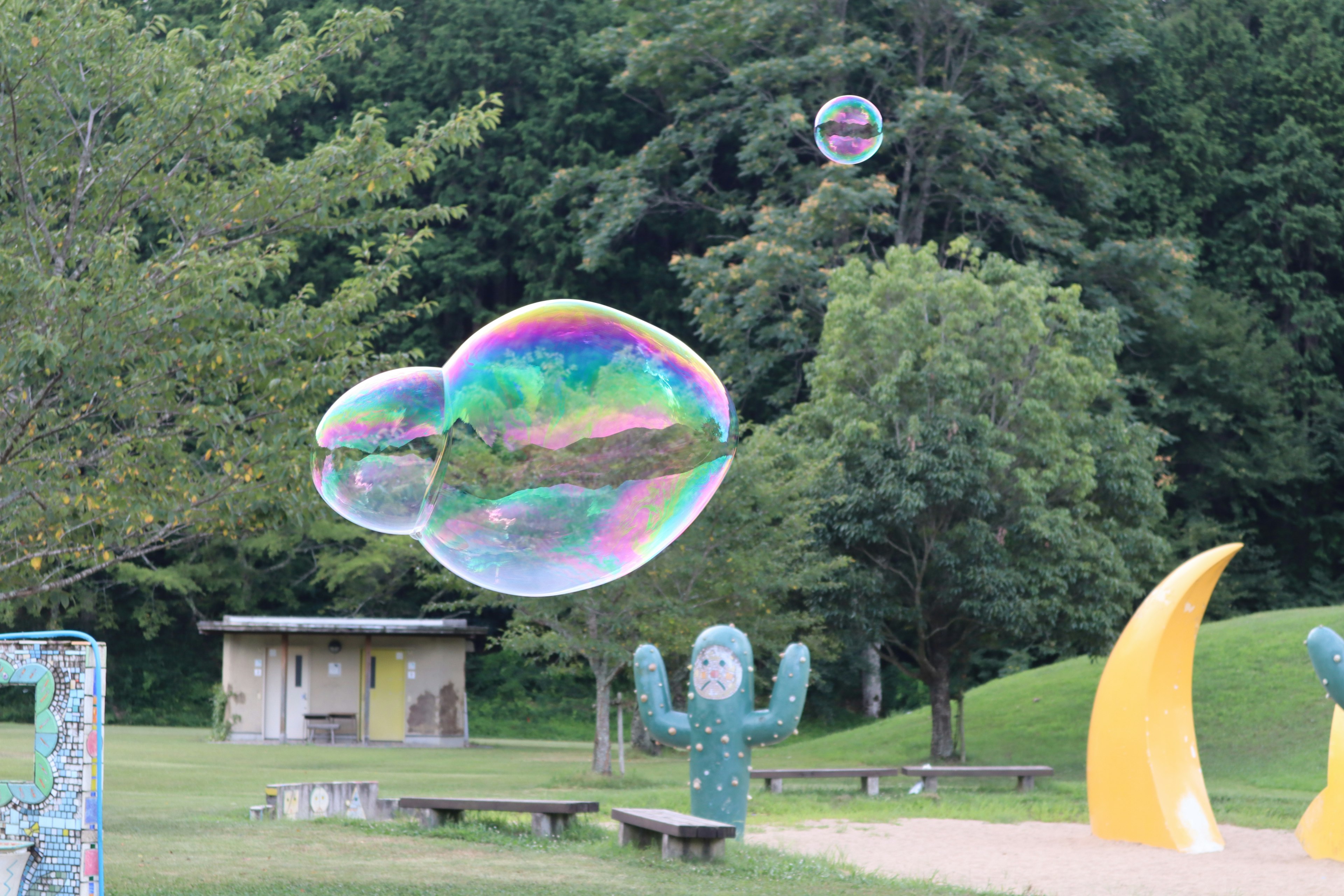 A large soap bubble floating in a park with green surroundings The bubble reflects rainbow colors