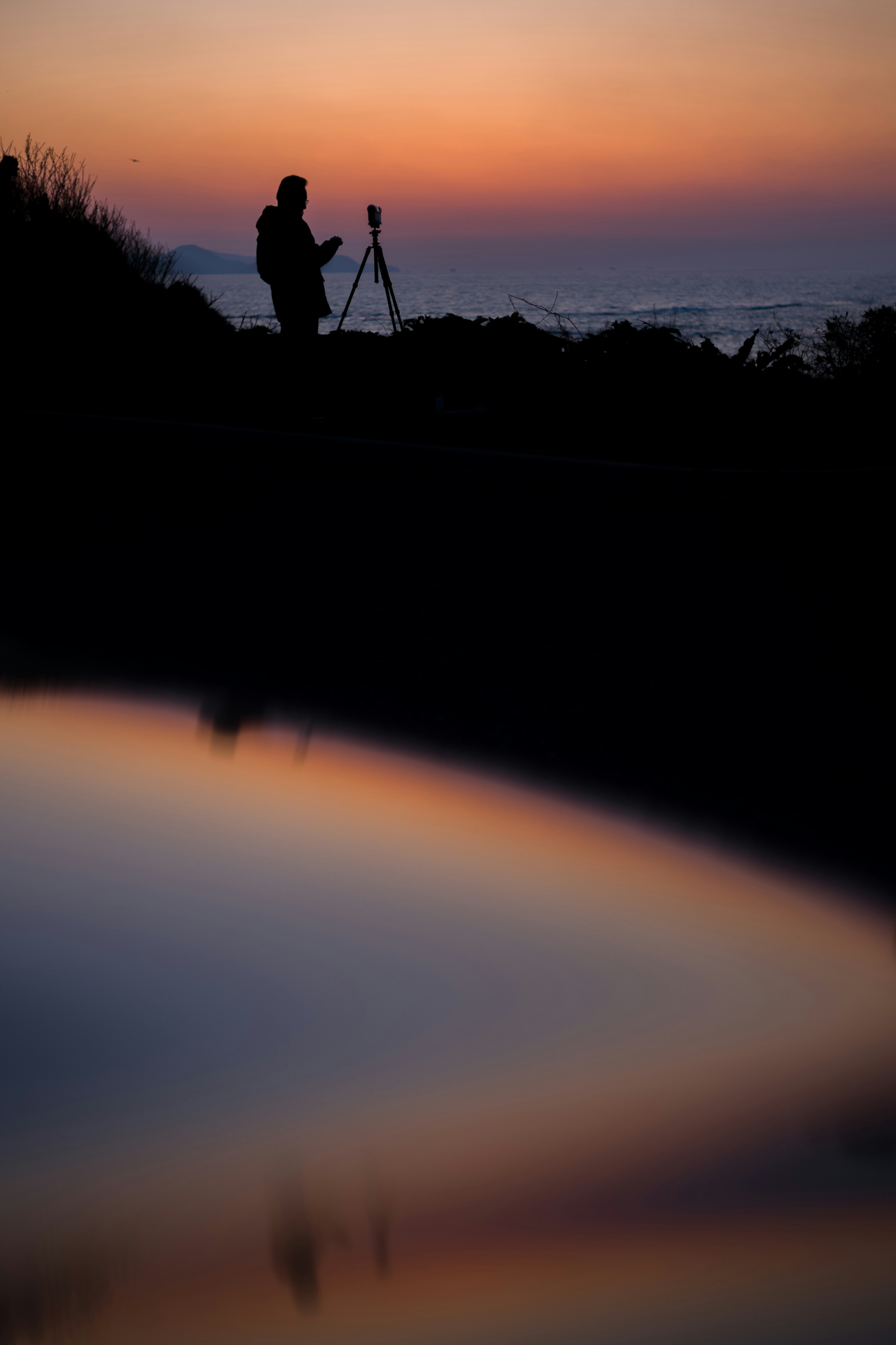 Silhouette di una persona con un treppiede al tramonto con riflessi sull'acqua