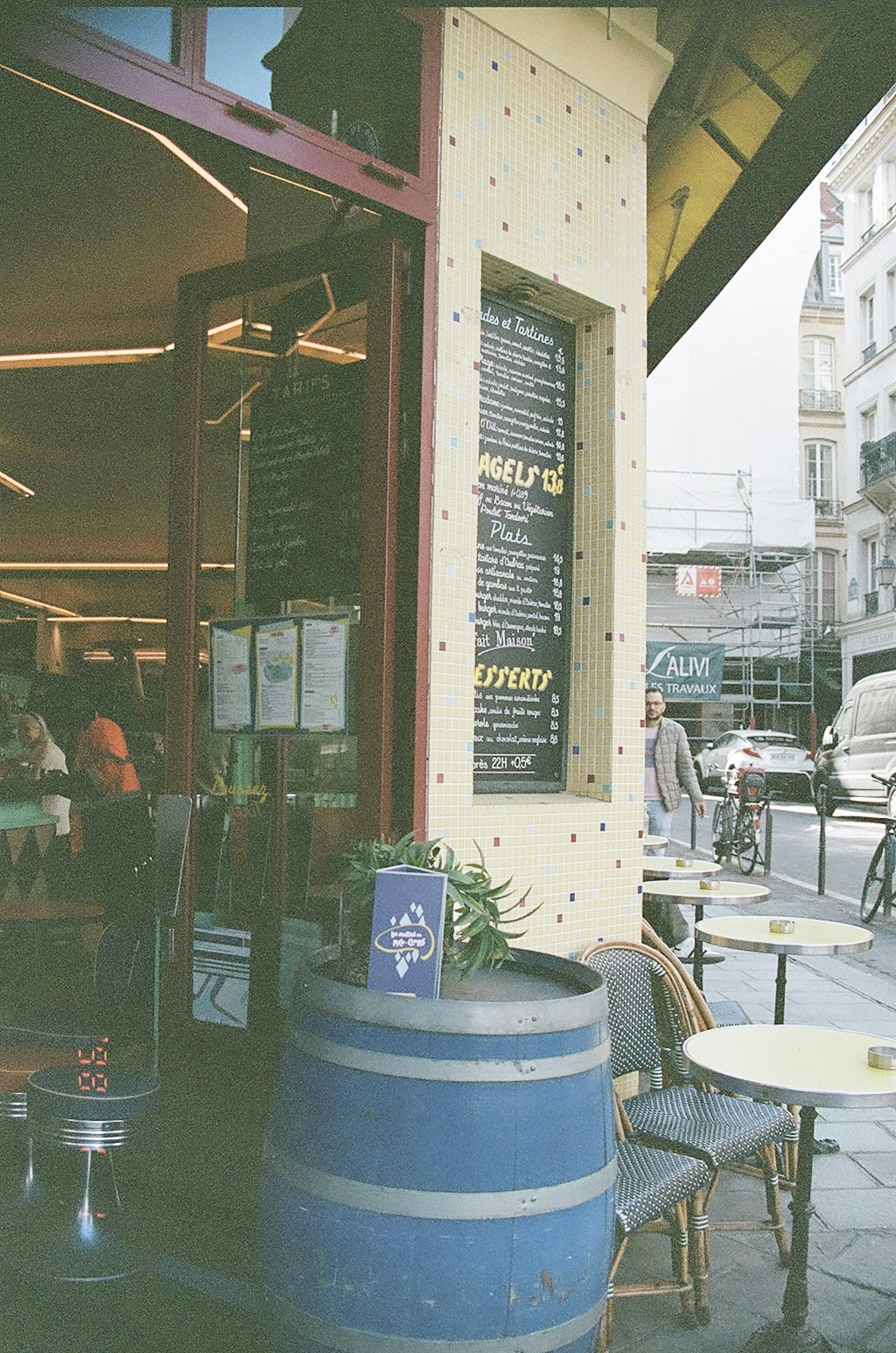 Eksterior kafe dengan barel biru, papan menu, meja dan kursi, suasana jalan yang hidup